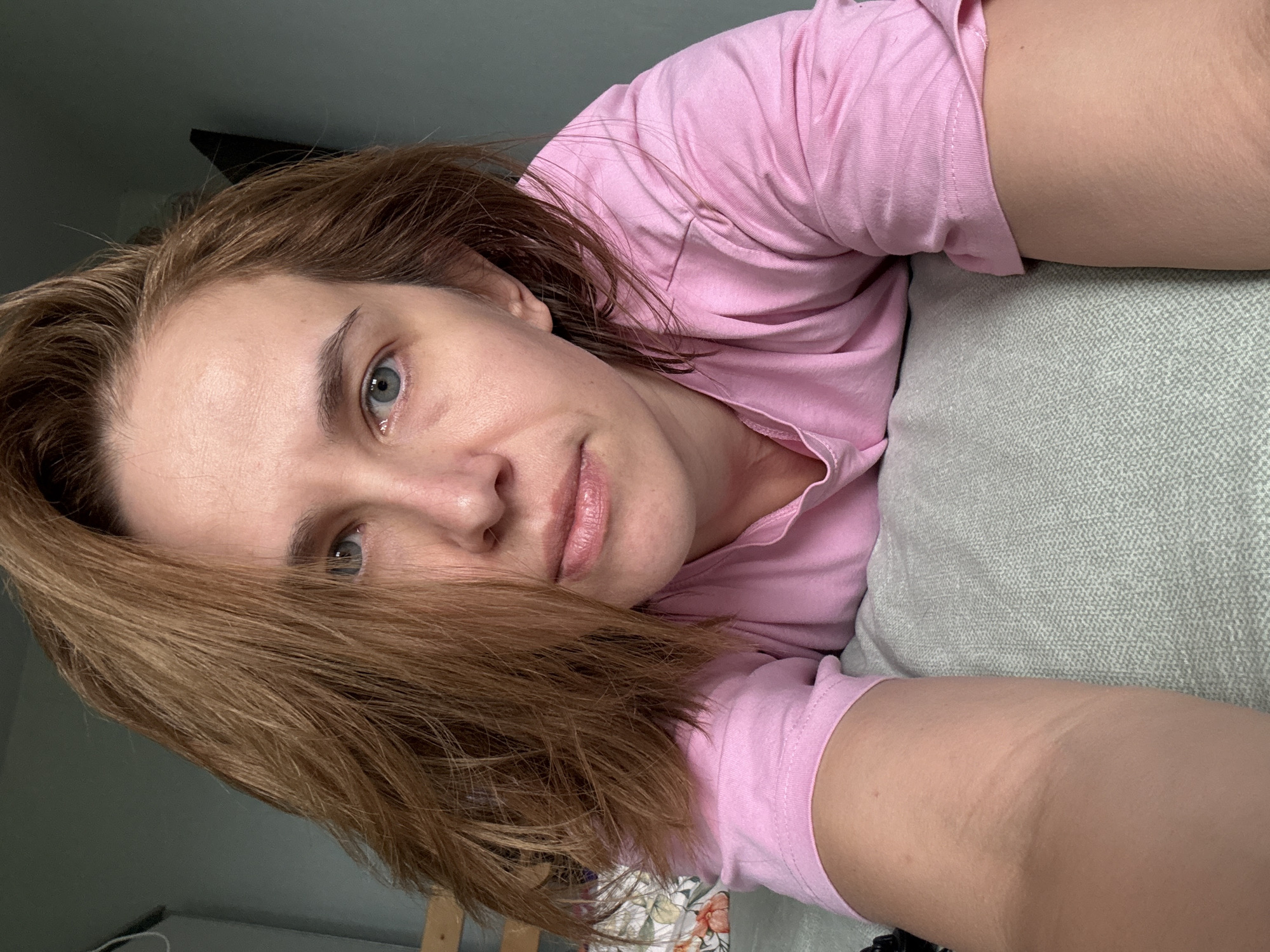 A woman with shoulder-length, reddish-brown hair, wearing a pink t-shirt, is lying on a gray surface that appears to be a bed. Her head is tilted forward, and she is looking towards the camera. Her eyes are blue and her lips are slightly parted. The background is a dimly lit room with a hint of a wooden structure on the left and a dark object on the wall to the right. There is no indication of the city of Moscow, Russia, within the image itself.