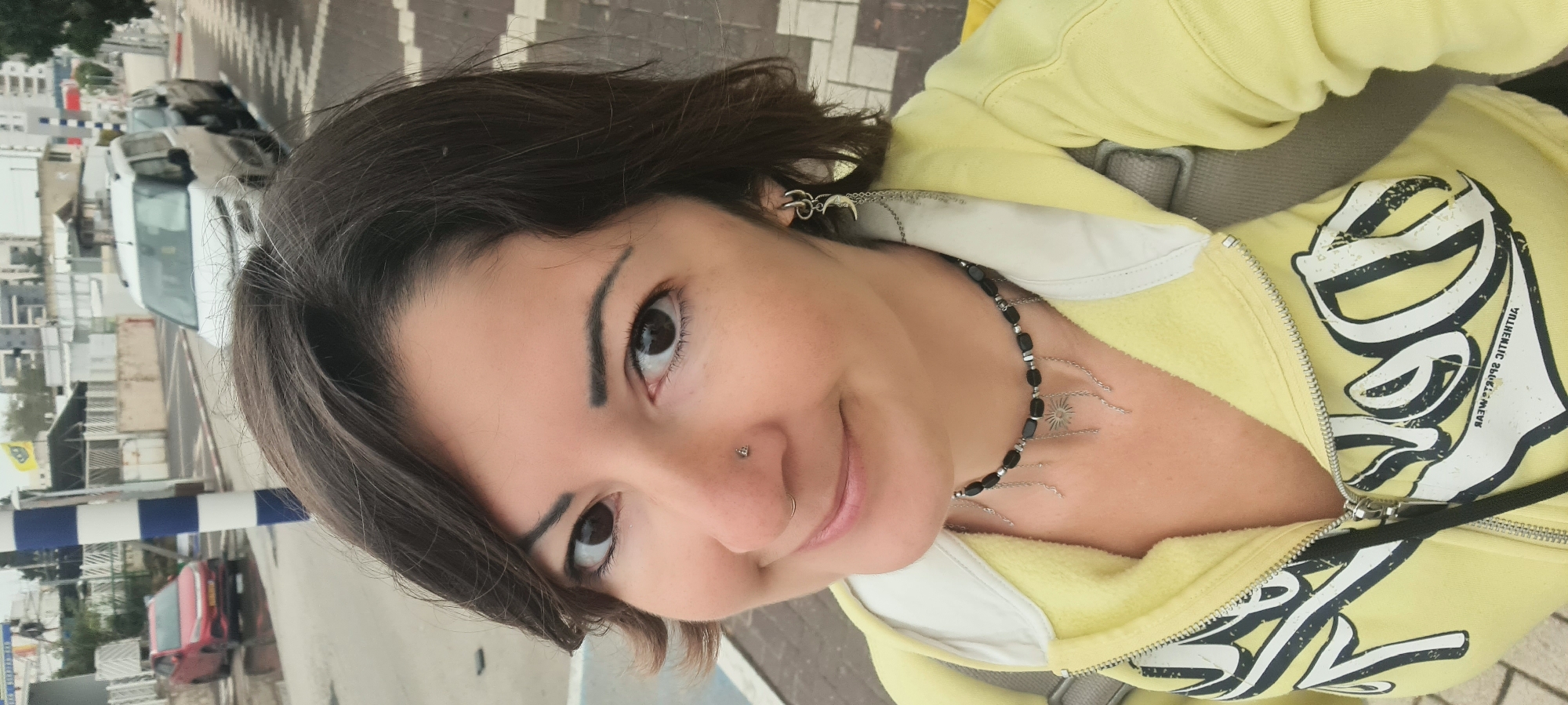 A woman is photographed close-up from a high angle. She has dark brown hair, dark eyes, fair skin, and a nose piercing. She is wearing a light yellow zip-up hoodie with a white collar and dark print. She is also wearing a black beaded necklace with silver charms and silver earrings. The background shows a city street, including parked cars, buildings, and a blue and white barrier. The location is Nahariyya, Israel.