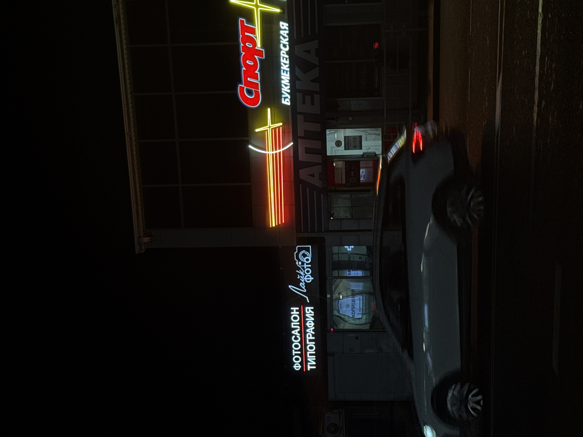 The image depicts a nighttime street scene in Abinsk, Russia. In the foreground, a white car is captured in motion, its headlights and taillights illuminated. Behind the car, several storefronts are visible, adorned with bright neon signs. One sign reads "СПОРТ" (Sport) in red and yellow, another, "БУКМЕКЕРСКАЯ" (Bookmaker's) in white, and below it, "АПТЕКА" (Pharmacy) in large grey letters. To the left, a sign for "ФОТОСАЛОН ТИПОГРАФИЯ" (Photo Salon Typography) is illuminated in white neon, with a stylized camera icon. The street itself is paved, and the buildings feature a modern, commercial architectural style, with large glass windows. The overall atmosphere is urban and commercial, illuminated by the artificial glow of the signs.