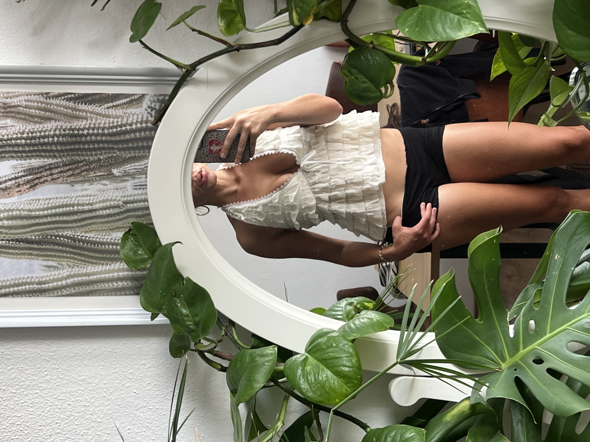 The image shows a reflection in a mirror of a person's upper body, arms, and legs. The person is wearing a white, ruffled, sleeveless top with lace trim and black shorts. They are holding a smartphone with a leopard print case in their right hand, angled towards the mirror. Their left hand is resting on their left hip. The person's lips are slightly parted, and they appear to be looking at their reflection.
The mirror is oval-shaped and framed in white. It is surrounded by lush green houseplants, with large, broad leaves that partially obscure the reflection and the surrounding room. In the background, visible in the reflection, is a framed artwork on a white wall, depicting a textured pattern of light and dark vertical stripes. Further in the background, a dark object, possibly a chair or bag, is visible, along with hints of other furniture and what looks like a small stuffed rabbit.
The lighting suggests an indoor setting, possibly during the daytime given the bright illumination. The overall atmosphere is casual and relaxed, with a focus on the person and their attire, presented within a natural, plant-filled environment. There is no visible text in the image, nor are there any specific location cues that tie it to Mühldorf a. Inn, Germany.
