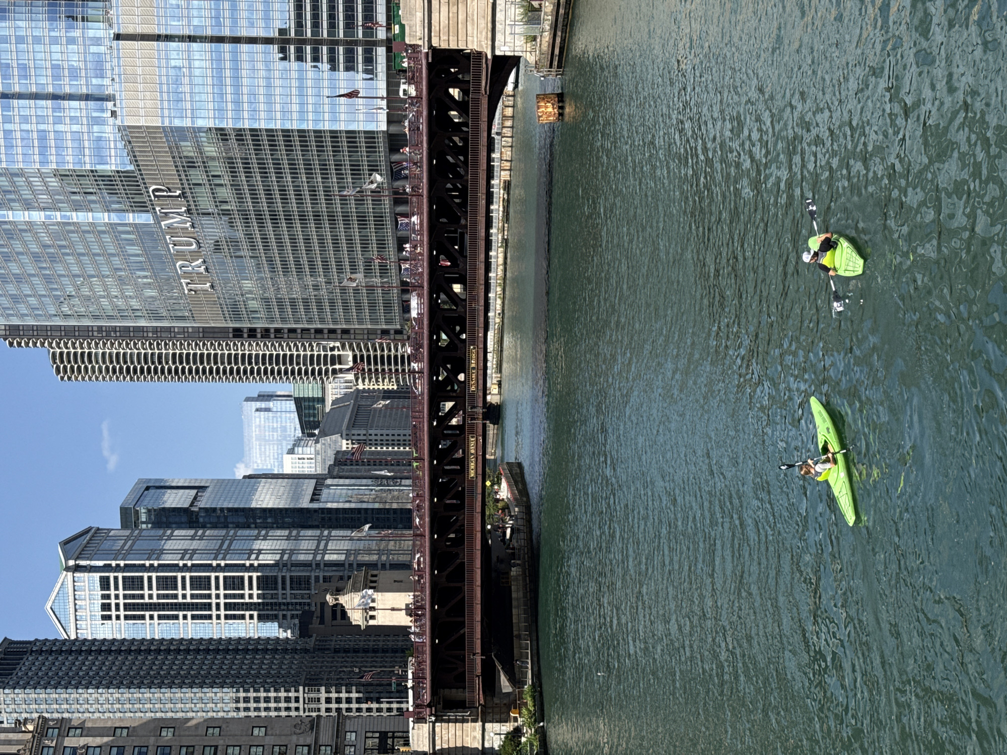 This media file captures a serene scene along the Chicago River on a clear, sunny day. The water, a calm greenish hue, reflects the surrounding urban landscape. Two individuals are seen kayaking, each in a bright green kayak, paddling leisurely. One kayaker is closer to the foreground, while the other is further in the distance, creating a sense of depth.
Dominating the left side of the frame is the iconic Trump International Hotel and Tower, its glass facade reflecting the blue sky and the building's grid-like structure. The word "TRUMP" is prominently displayed in large, silver lettering on the building. Adjacent to the skyscraper, a metal bridge, likely a bascule bridge, spans the river. The bridge is intricate in its design, with visible metalwork and supports. People can be seen walking and standing on the bridge, enjoying the view. American flags are also visible on the bridge and buildings, adding a patriotic element.
Further down the river, more modern skyscrapers with glass exteriors punctuate the skyline, showcasing Chicago's architectural grandeur. The overall impression is one of a vibrant city with a thriving riverfront, where urban life and recreational activities coexist harmoniously. The lighting suggests it is daytime, likely late morning or early afternoon, given the bright sun and clear visibility. The weather appears pleasant, conducive to outdoor activities like kayaking and sightseeing. Text visible on the bridge reads "MICHIGAN AVENUE" and "DU SAU" which is part of "DuSable Bridge".