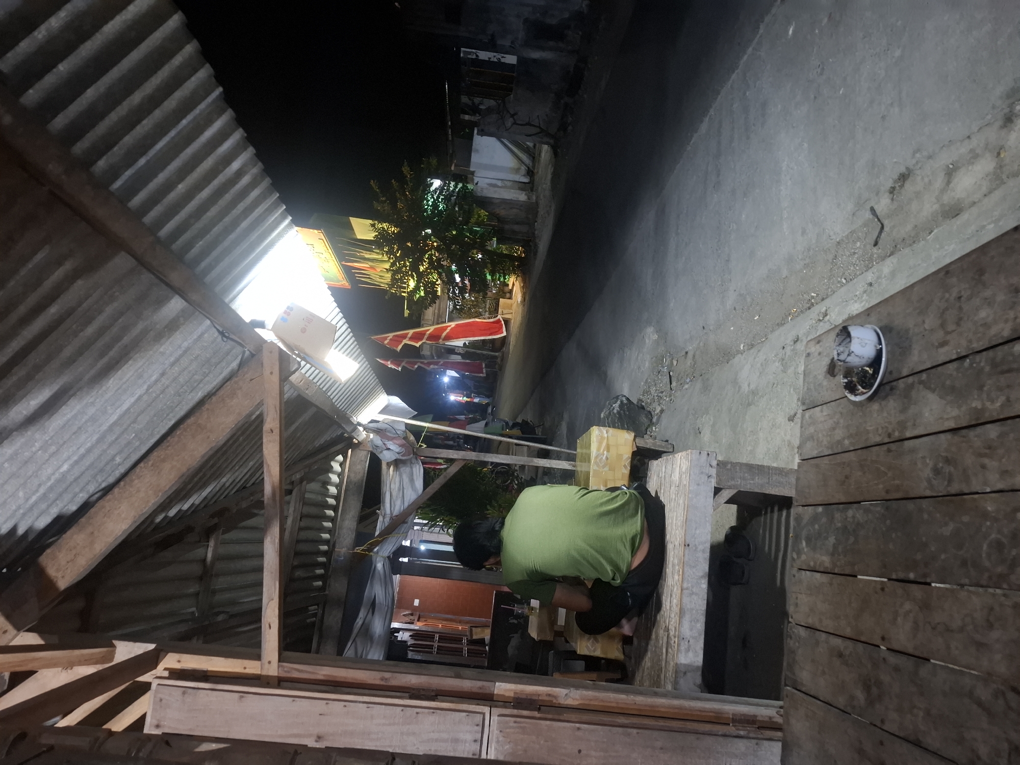 An exterior, low-angle shot captures a dimly lit scene at night along a paved road in Bulubrangsi, Indonesia. In the foreground, a person wearing a green t-shirt and dark pants sits on a wooden bench, facing away from the camera. They appear to be in a covered outdoor seating area with a corrugated metal roof supported by wooden beams. To the left, a wooden structure with vertical planks is visible. In front of the person, there is a low wooden table. To the right of the seating area, the paved road stretches into the distance, illuminated by ambient light from nearby structures. Red and white flags are displayed on poles along the side of the road, and a tree with artificial lighting is visible behind them. The ground surface is a mix of concrete and loose gravel. In the immediate foreground, on a wooden plank surface, rests a small, white ashtray filled with ash and cigarette butts, situated on a white plate. The overall atmosphere is that of an informal roadside establishment at night.