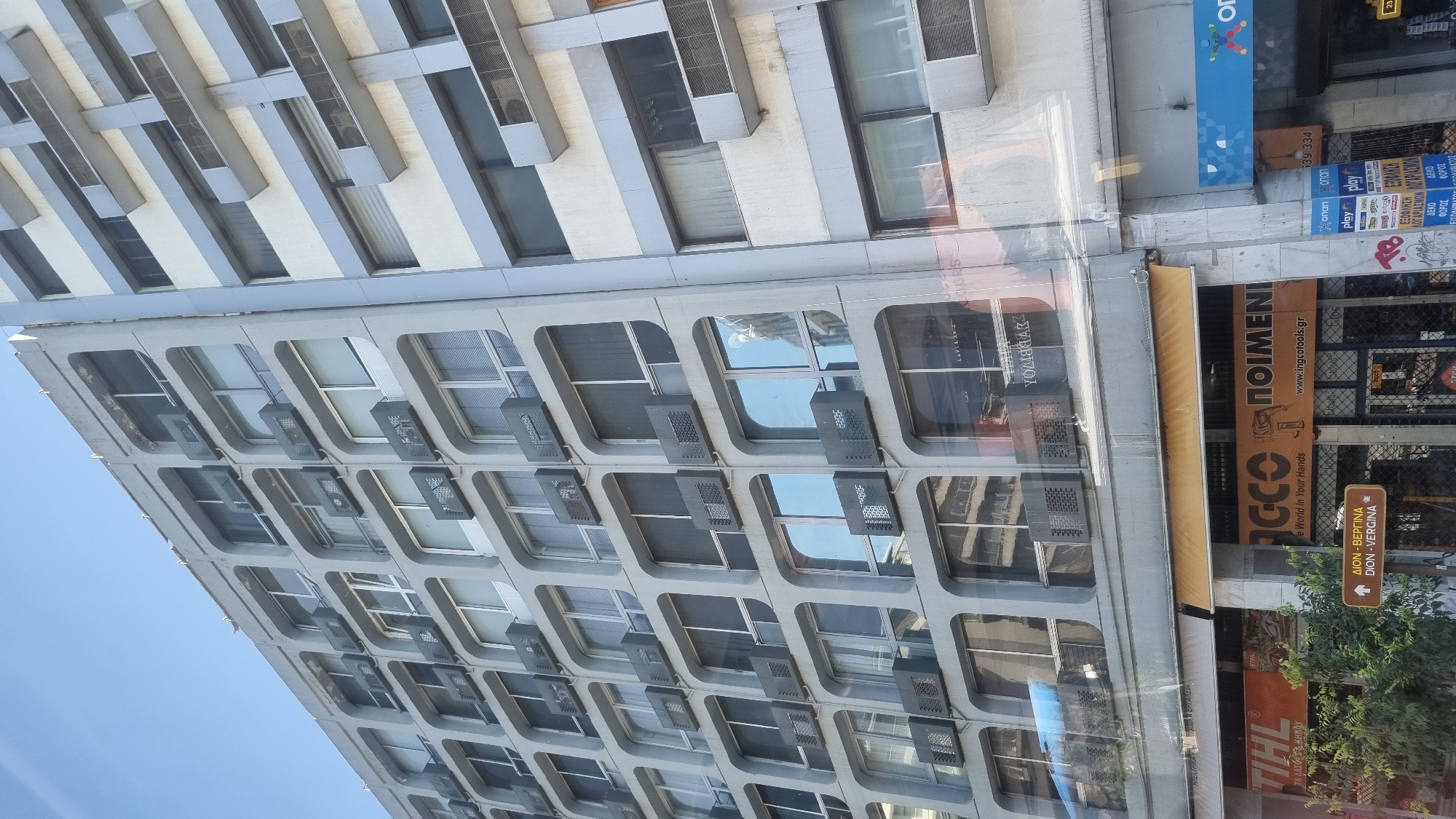 This is a full, eye-level, slightly upward-angled, medium shot of a concrete apartment building in Thessaloniki Municipal Unit, Greece. The building is light grey and has a grid-like facade with numerous windows and air conditioning units. The windows are mostly covered with blinds or curtains. The building is tall and appears to be in a densely populated urban area, as suggested by the presence of other buildings visible in the background and the street-level businesses. The sky is clear and blue. The street-level features include awnings, shop signs, and trees. One sign reads "STIHL," another has "YOMBARAK" visible in reflection on the windows, and another reads "POIMENI." Another sign in Greek indicates directions to "DION-VERGINA." The overall impression is one of a typical urban street scene in Greece.