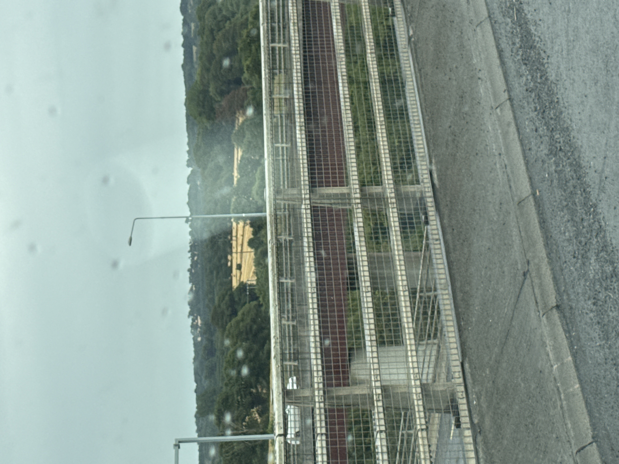 The image captures a view from an elevated position, likely a bridge or overpass, looking out over a rural landscape under an overcast sky. In the foreground, a concrete road surface with a curb is visible, suggesting the perspective is from a vehicle or walkway. Beyond this, a metal fence, likely a safety barrier, runs horizontally across the frame, composed of vertical posts and horizontal railings with a grid-like mesh.
Beyond the fence, a multi-tiered structure is discernible, possibly another level of the same overpass or an adjacent building. This structure features dark red rectangular panels and a lighter colored grid pattern. Further in the background, a dense line of trees forms a verdant horizon. Within this treeline, glimpses of man-made structures and fields can be seen. One field appears to have been recently harvested, with a pattern of straw bales or stubble visible.
A solitary street lamp with a curved arm is present in the mid-ground, reaching above the fence line. The overall atmosphere is muted due to the overcast weather, with a slightly hazy quality, possibly due to rain or moisture on the camera lens, evidenced by several circular distortions throughout the image. The location is identified as being in the city of Grannagh, Ireland.