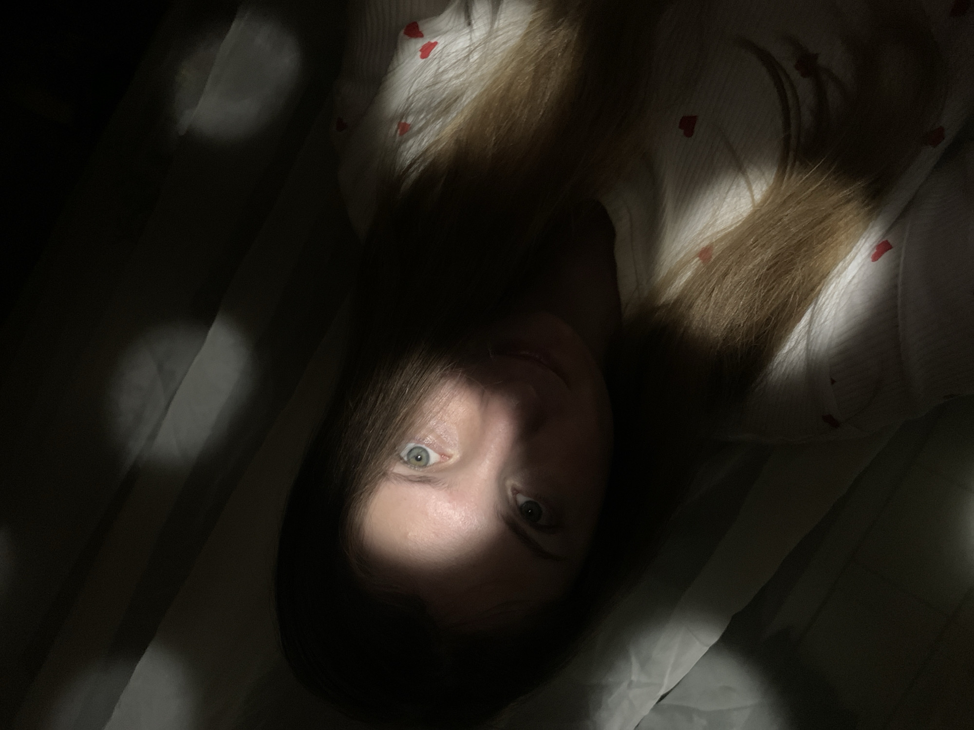 An intimate, dimly lit portrait features a person, likely a woman, lying down in what appears to be a bedroom in Chaikovsky, Russia. The image is taken from an overhead perspective, with the person's head slightly tilted. Long, light brown hair is spread around their head on a light-colored surface.
The most striking aspect of the image is the dramatic lighting: a strong, narrow beam of light illuminates the left side of the person's face (viewer's left), particularly highlighting a wide, alert, bluish-green eye. The right side of the face and the majority of the room are cast in deep shadow, creating a stark contrast and a mysterious ambiance. The other eye is mostly obscured by shadow, though it appears open. The person is looking directly towards the camera, their visible eye conveying a sense of direct engagement or possibly surprise.
The person is wearing a white garment, possibly nightwear or a top, adorned with small red heart patterns, visible on the upper body and within the lighter areas of the image. Below the person, a striped fabric, possibly a bedsheet or blanket, is partially visible in the shadows and caught by incidental light. The overall darkness suggests it is either very early morning or late evening. There is no visible text in the image.