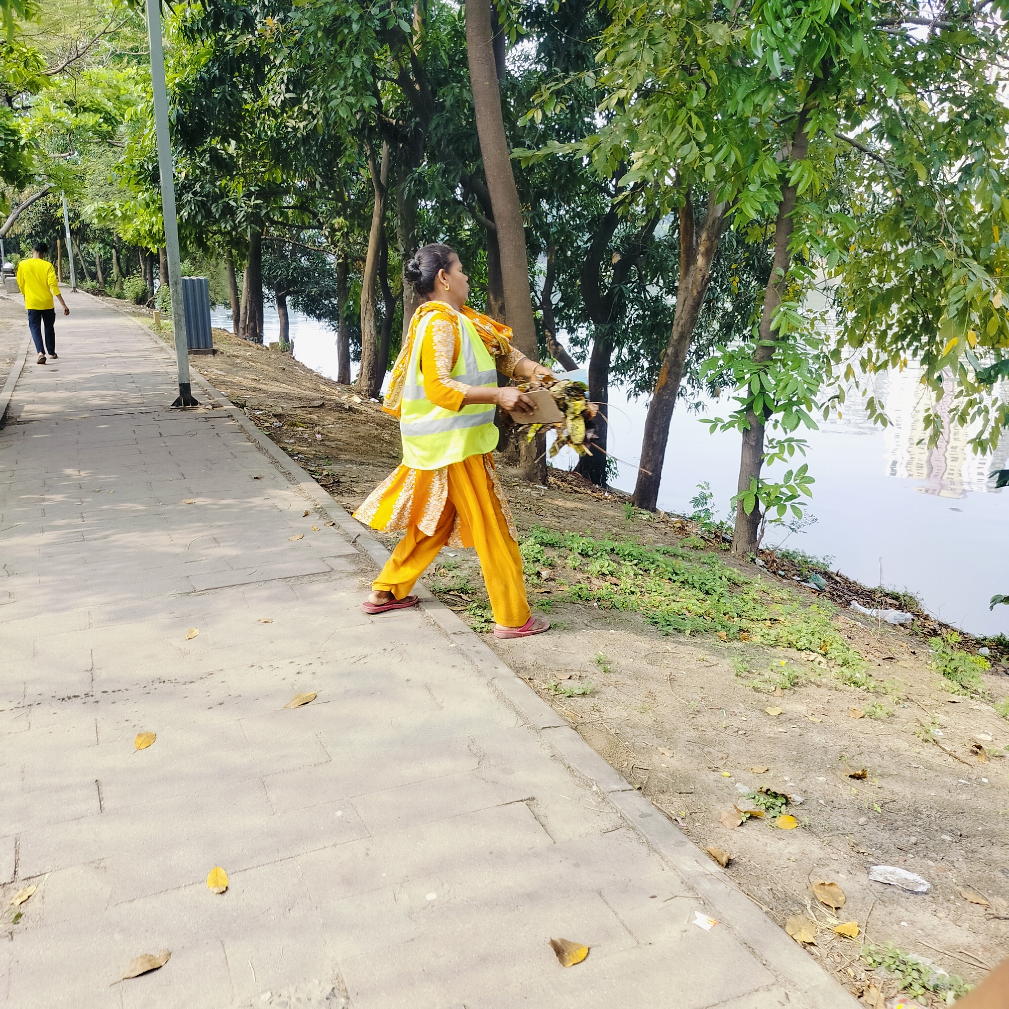 A woman wearing an orange salwar kameez with a yellow high-visibility vest is collecting fallen leaves and debris from the edge of a paved walkway near a body of water. She is carrying the gathered waste in her hands. The walkway borders a grassy slope leading down to the water's edge, which has trees along it. Litter is also present on the grass. Across the water, building reflections are visible. In the background, on the walkway, a person in a yellow shirt walks away from the camera. The scene depicts a park or recreational area in Dhaka, Bangladesh.