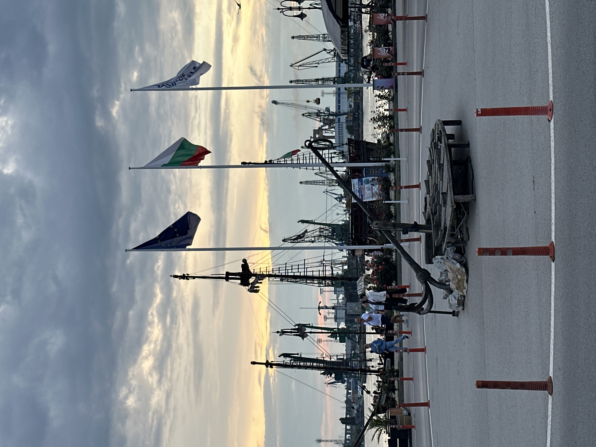 The image captures a vibrant port scene in Varna, Bulgaria, at what appears to be sunset or sunrise. The sky features a dramatic blend of warm orange and yellow hues near the horizon, transitioning into cooler grey and blue clouds overhead.
In the foreground, a paved area is visible, bordered by several orange traffic bollards. Three flags fly prominently on tall flagpoles against the sky: the national flag of Bulgaria (white, green, red horizontal stripes), the flag of the European Union (blue with a circle of gold stars), and a white flag with blue text that likely reads "PORT OF VARNA," with "OF" and parts of other letters clearly discernible. Near the paved area, a large, antique ship's anchor is displayed on a bed of rocks, next to what appears to be a segment of an old wooden cart or wagon wheel.
Several people are scattered throughout the scene, casually walking or standing, suggesting a public promenade or an event. One person in a light blue outfit is particularly noticeable in the mid-ground. The area along the waterfront features decorative elements and display stands, hinting at a public gathering or a tourist-friendly spot.
The background is dominated by the industrial infrastructure of the port. Numerous large gantry cranes are silhouetted against the illuminated sky, towering over what appears to be water and other port buildings. Masts and superstructures of several docked ships or large boats are also visible, further emphasizing the port setting. The overall atmosphere is calm and expansive, with the fading or rising sunlight casting long shadows and highlighting the outlines of the port's machinery.