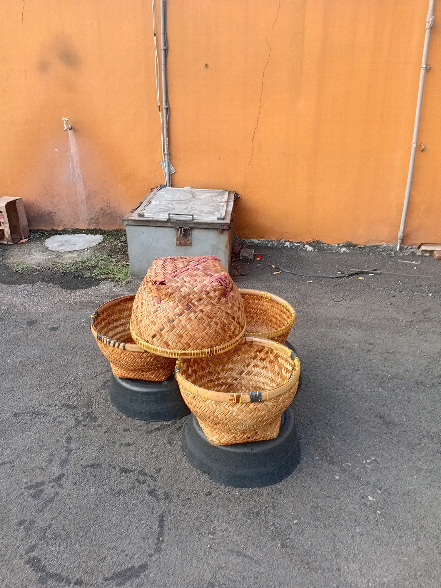 Four woven bamboo baskets are stacked on two black circular bases, likely repurposed tires. The baskets vary in size and are arranged in a tiered fashion, with one large basket on top, two medium-sized baskets underneath, and one smaller basket at the bottom front. The scene is set against an orange wall with a crack running vertically down its center. To the left of the baskets, a water tap is visible, with water spraying out. Behind the baskets, a metal storage box with a lid is present. The ground is paved with asphalt. The overall environment suggests an outdoor space, possibly a backyard or alleyway, in Tangerang, Indonesia, as indicated by the context provided. There are no people visible in the image.