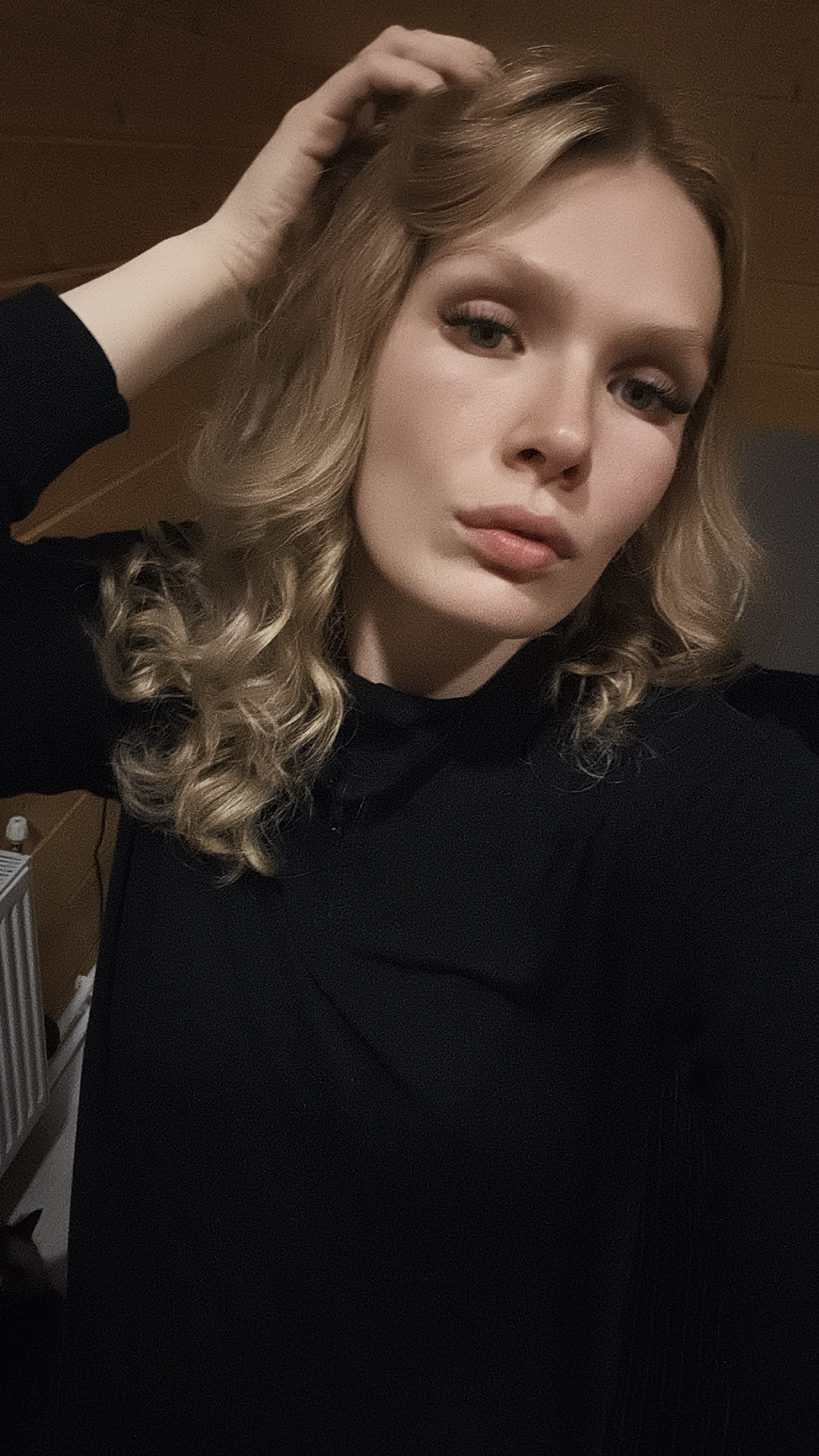 A woman is shown in a close-up shot. She has blonde hair with curls and is wearing a black top. Her hand is raised, touching her hair. The lighting is dim. A radiator is faintly visible in the lower left corner, and wood paneling is visible in the upper background. The location, Русский Вожой, Russia, cannot be determined from the image.