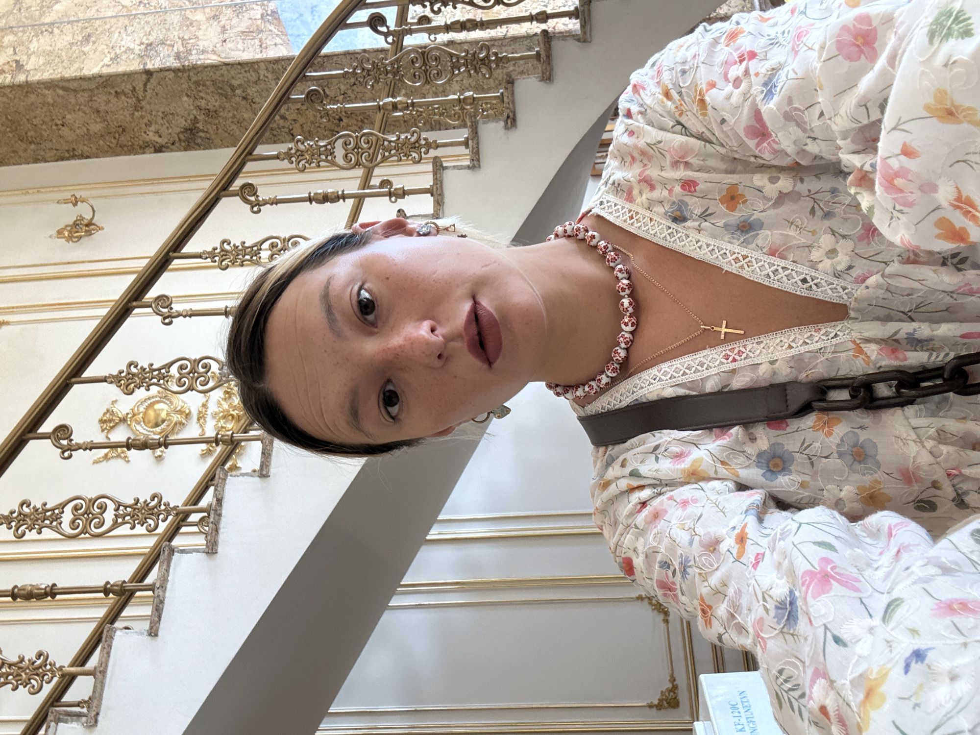 A woman with brown hair pulled back, a pale complexion with freckles, and dark eyes takes a selfie in front of an ornate staircase. She is wearing a white, floral-print dress with a deep V-neck and is adorned with a beaded necklace and a gold cross pendant. A dark bag with a chunky chain strap is slung over her shoulder. The staircase features gold railings with intricate scrolling metalwork and marble steps, suggesting a grand interior. A portion of a blue sign with text in Vietnamese is visible in the lower left corner, with "KF-120C" and "NGFUNET.VN" discernable, indicating a location in Vietnam. The overall setting appears to be a well-decorated indoor space, possibly a hotel or a public building in Đà Nẵng.