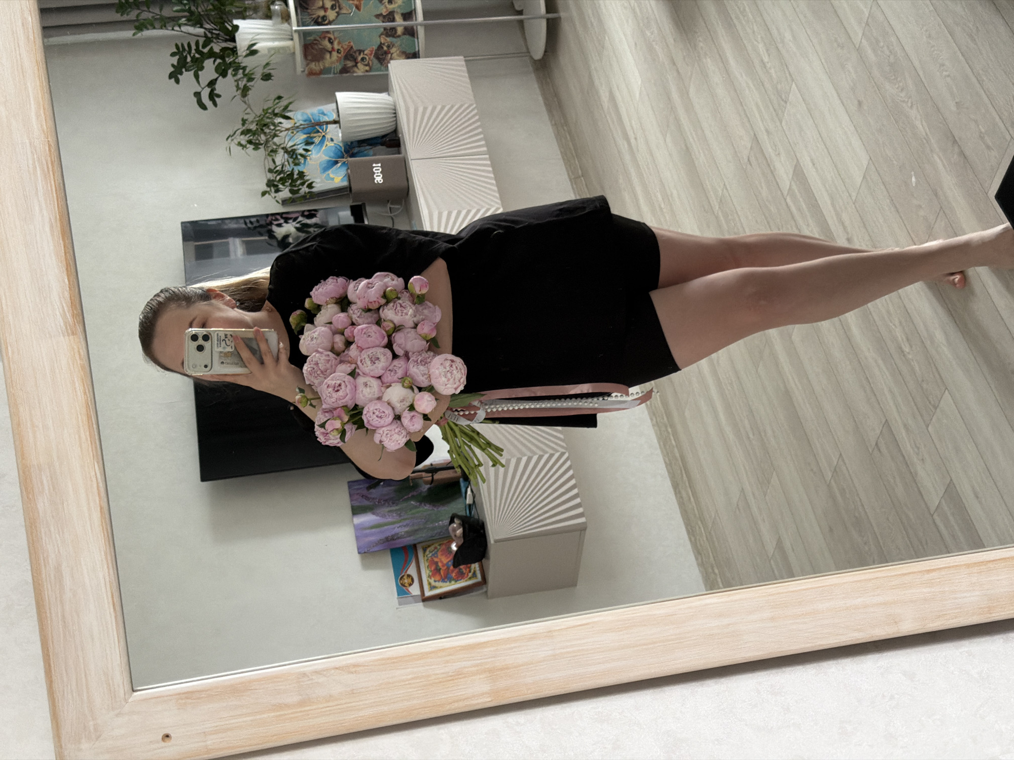 The image presents a mirror selfie of a woman, seen from the mid-chest down, holding a substantial bouquet of tightly packed light pink flowers, consistent with peonies or garden roses. Her face is obscured by the smartphone she holds to take the photograph. She has light-colored hair pulled back in a ponytail and is wearing a loose black short-sleeved top and black shorts. The flower stems are tied with a pink ribbon and decorated with a string of white beads or pearls. She stands barefoot on a light-colored wooden or laminate floor.
The room reflected in the mirror features light-colored, textured walls. To the left of the woman, a light-colored floating shelf with a distinctive radial sunburst pattern holds a small black bowl containing light-colored items, a framed image of flowers (possibly poppies), and another framed artwork depicting a lavender field. To her right, a low, white console table, also with a radial sunburst pattern, displays a digital clock showing "00:01," a white potted plant, and a larger green plant in a white pot. A large flat-screen television is visible behind the console, reflecting parts of the room. Further to the right, a tall white shelving unit is adorned with multiple green potted plants and decorative items, including small artworks featuring cartoon cats. The setting is an indoor domestic environment. The image contains no visual information or identifying markers to specify its location within the city of Chaikovsky, Russia.
