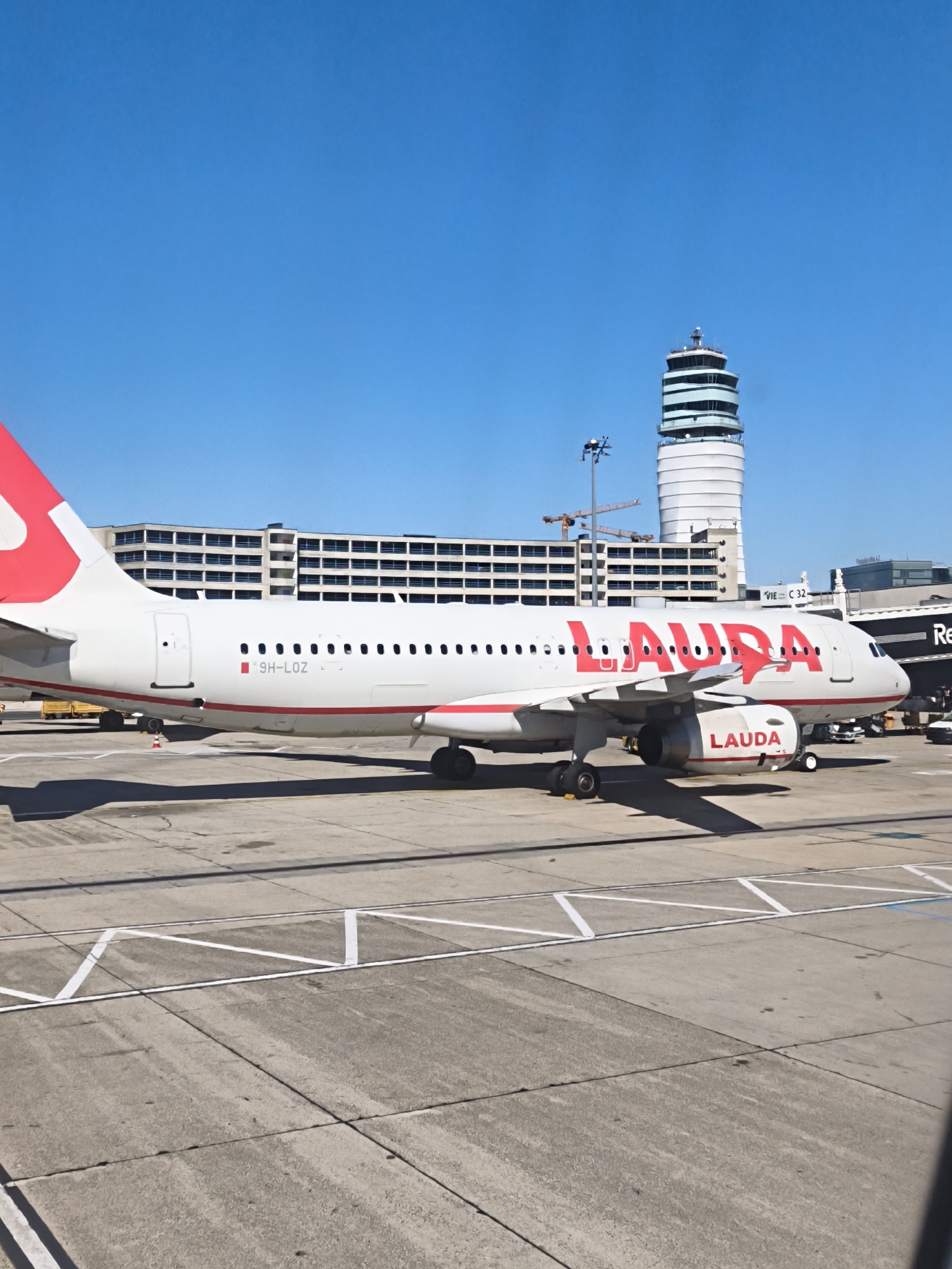 The image shows a Lauda airplane parked on the tarmac at Vienna International Airport (VIE) in Schwechat, Austria. The airplane is white with red lettering that spells out "LAUDA" on its side and red accents on its tail and wingtips. A Lauda logo is also visible on the engine. In the background, a modern airport terminal building and a control tower are visible against a clear blue sky. Several other airport vehicles and structures are present, including what appears to be a jet bridge attached to the terminal. The ground is paved with concrete and marked with white lines for taxiing and parking. The scene suggests a typical operational day at a busy international airport.