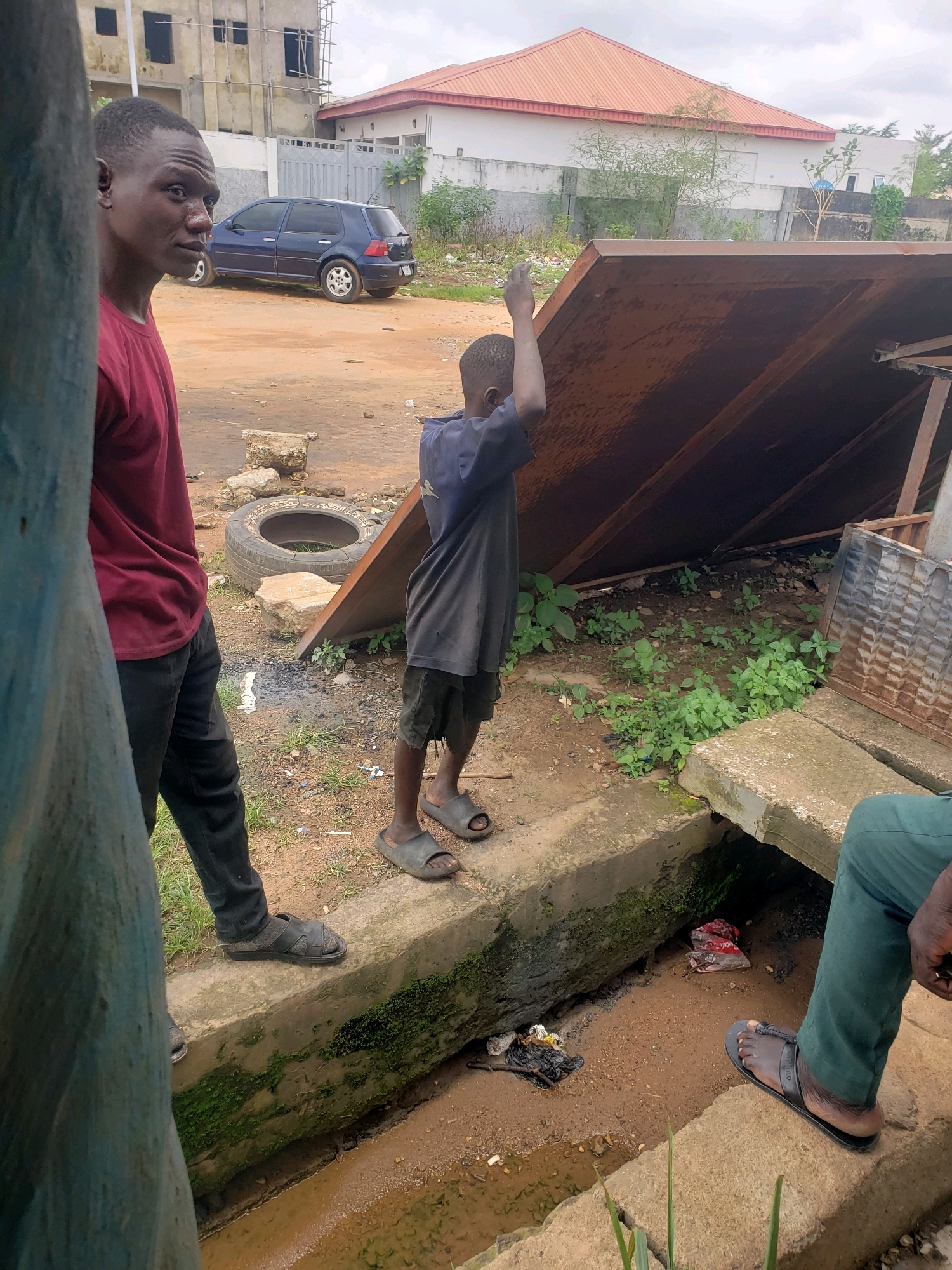 In Sabon Tasha, Nigeria, a group of people are gathered outdoors. A young boy, wearing a dark t-shirt and shorts, stands with his back to the camera, raising his right hand as if to signal or push an object. He is barefoot, wearing dark sandals. To his left, a young man in a maroon t-shirt and black pants looks to his left, his expression somewhat obscured. He is also wearing black sandals. In the background, a blue hatchback car is parked on a dirt road in front of buildings. A large, rusty metal sheet leans against a structure, partially covering a patch of green vegetation. The ground is sandy and littered with debris, including a discarded tire and concrete blocks. A concrete drainage channel runs along the foreground, containing muddy water and trash. Another person, wearing green pants and dark sandals, is visible in the lower right corner, standing near the drainage.