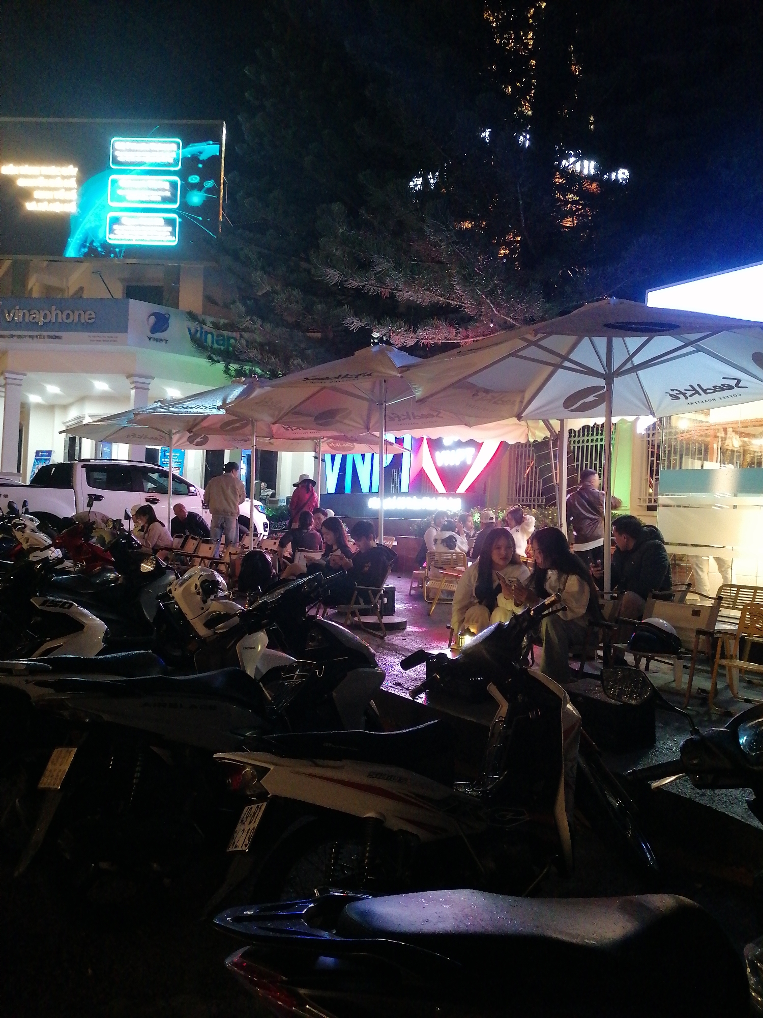A nighttime street scene in Da Lat, Vietnam shows an outdoor cafe setting. Several parked motorbikes dominate the foreground. Customers are seated at tables under large umbrellas, enjoying food and drink. A Vinaphone store is visible in the background, as well as a large LED display.