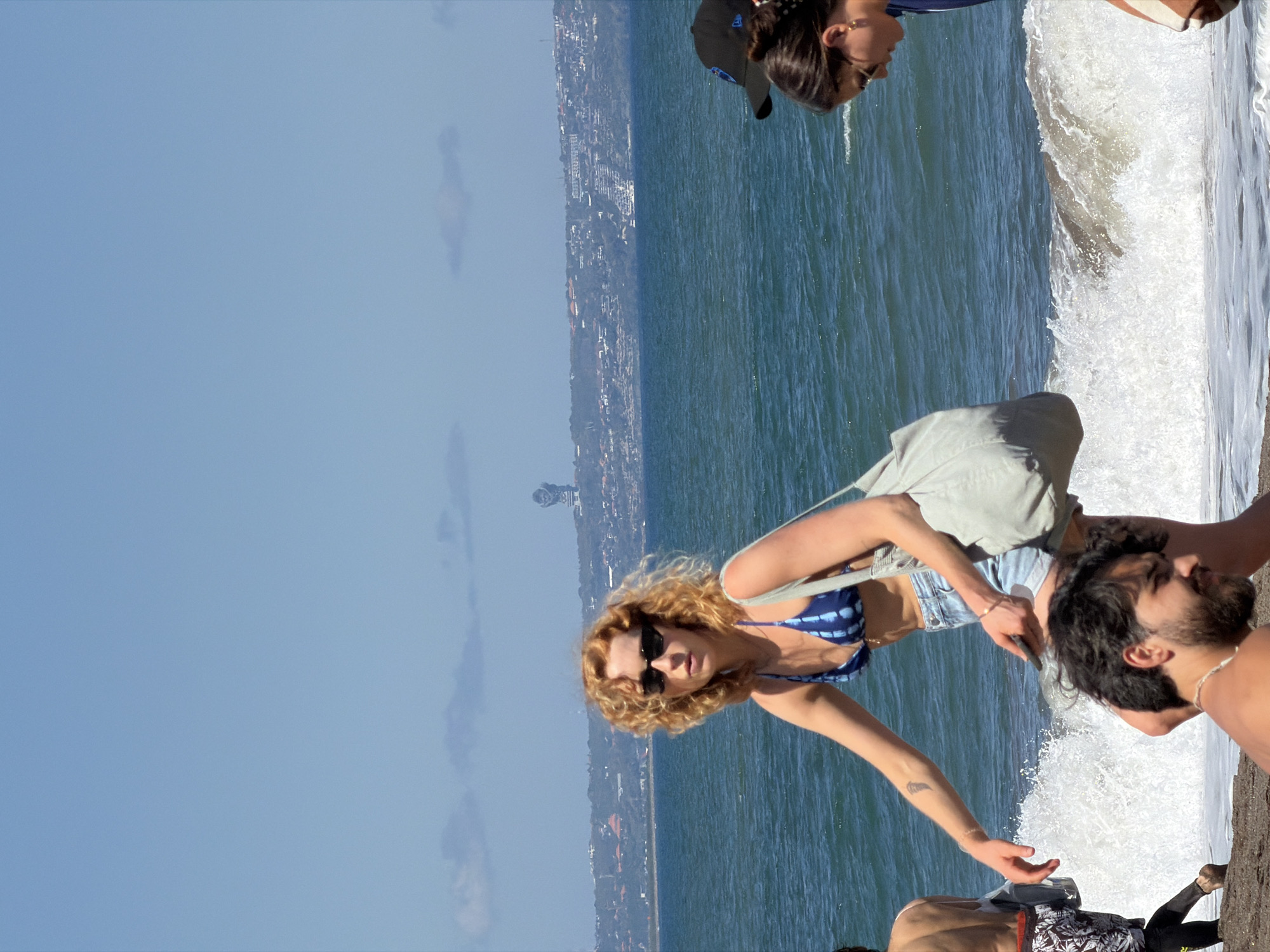 The image shows a group of people on a beach in North Kuta, Indonesia. A woman with curly blonde hair and sunglasses, wearing a blue and white bikini top and denim shorts, stands near the shoreline. She is reaching towards the water. Next to her is a man with dark hair and a beard, standing close to the waves. Behind them, another individual wearing a baseball cap is partially visible. The background includes the ocean and a view of the urban landscape of North Kuta. The sky is blue with some clouds. Waves are crashing on the shore. Another partially visible person is wearing what looks like dark black pants and a black and white top with a hint of red on the side.