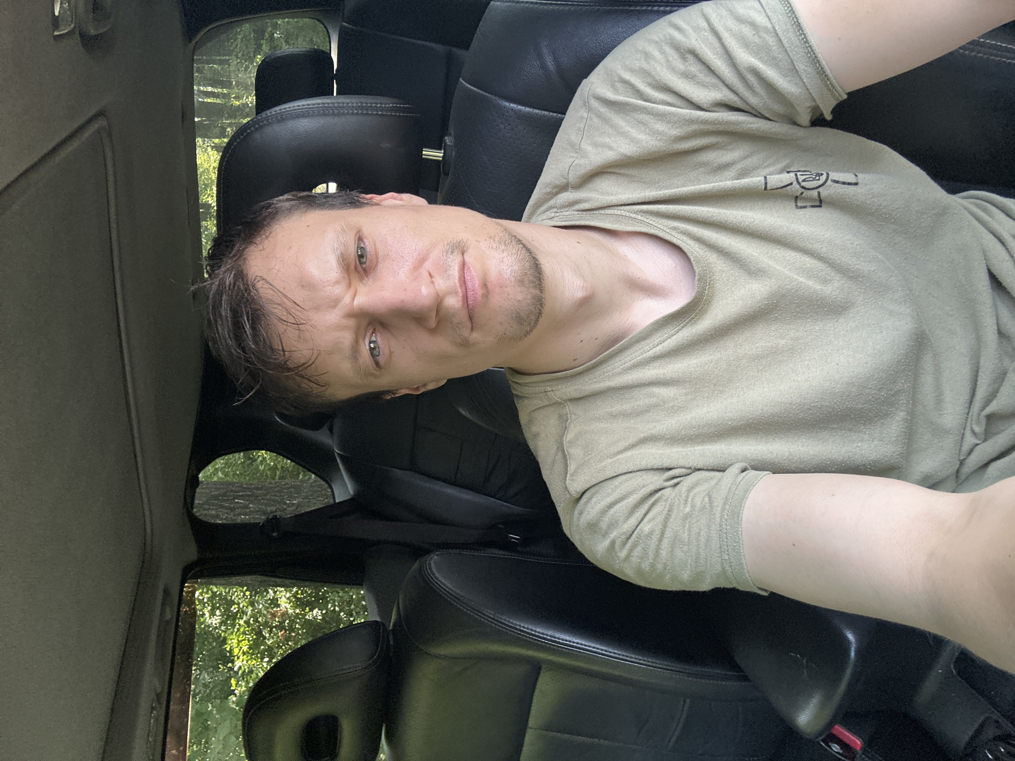 A man is sitting in the driver's seat of a vehicle, looking directly at the camera. He has short, dark hair and a light complexion. He is wearing a light brown t-shirt with a graphic on the left side of the chest. The interior of the vehicle is visible, with black leather seats and the roof liner. Through the windows, trees and foliage are visible, suggesting an outdoor, possibly rural, setting. The image appears to be a selfie. The location is Makiv, Ukraine.