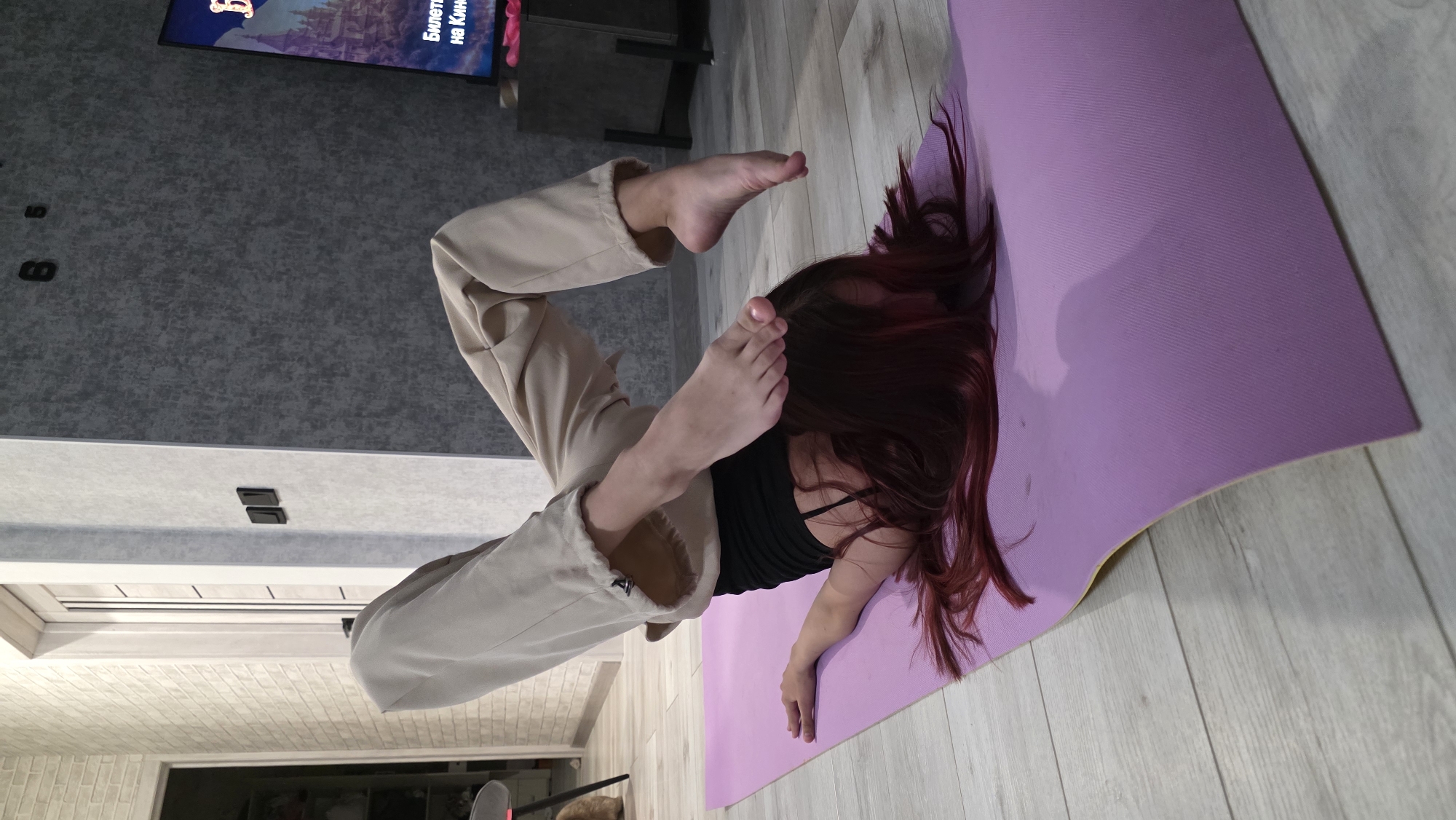 A woman is performing a yoga pose on a purple yoga mat in what appears to be an indoor setting. She is wearing a black tank top and beige pants, and her hair is dark red/maroon, covering a portion of the mat. Her legs are contorted, with her feet near her head. The room has wood-patterned flooring. A television is visible in the background displaying an animation, with a gray wall and a dark gray cabinet underneath the television. No specific location information, such as the city of Bulgakovo, Russia, can be determined from the image.