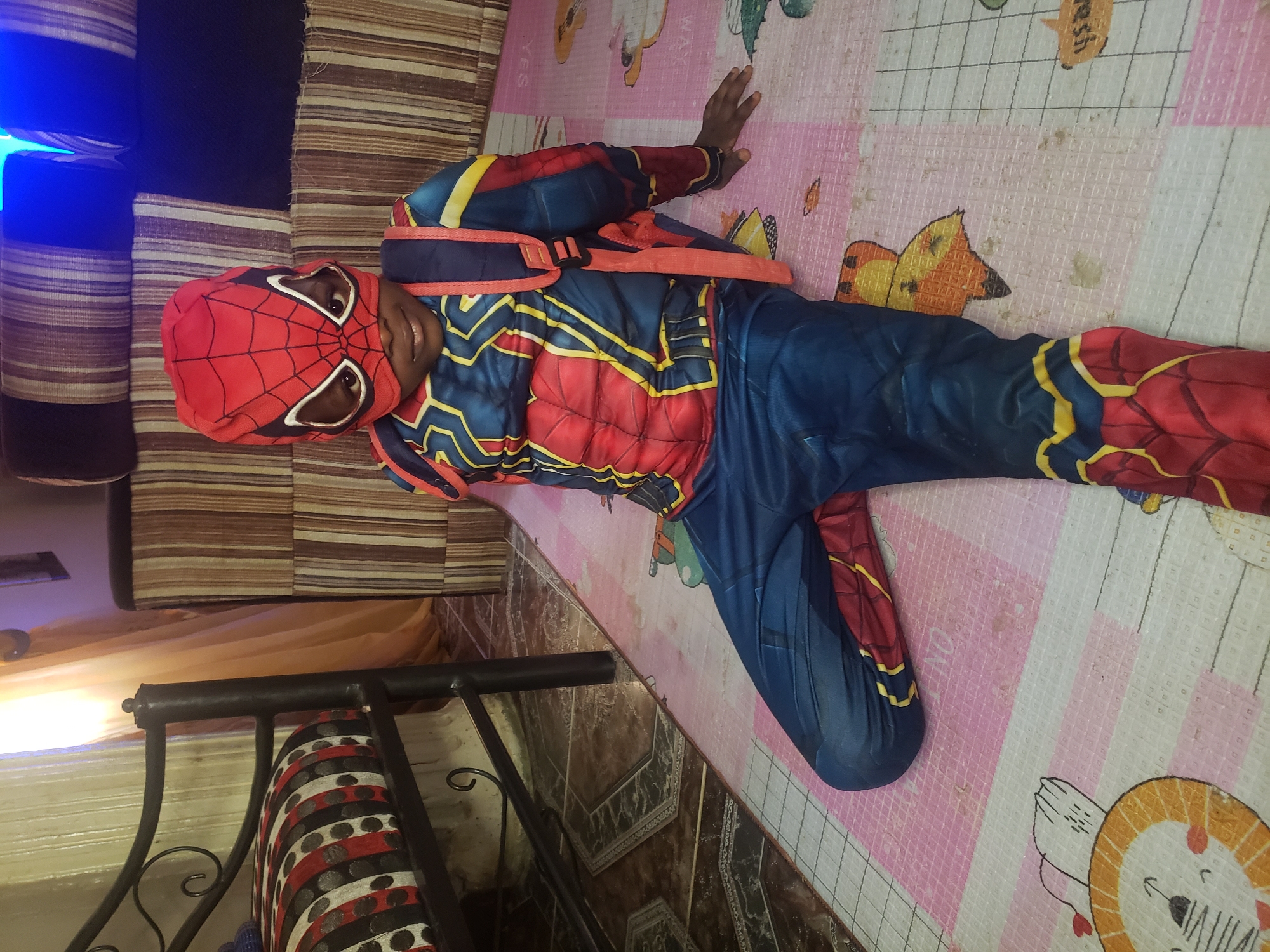In Narayi, Nigeria, a young boy dressed as Spider-Man sits on a patterned mat on the floor, leaning against a striped couch. He is wearing a full Spider-Man costume, including a mask that covers his head and face, with white eye openings. The costume is predominantly red and blue, with yellow accents and a web design. A blue backpack with red straps is worn over the costume. The room appears to be a living area, with a metal-framed bed with a patterned mattress visible to the left, and a wooden tiled floor partially covered by the mat. The lighting in the room suggests it is daytime, with natural light coming from a window or opening behind the couch, and artificial blue light originating from somewhere above and to the right of the frame.