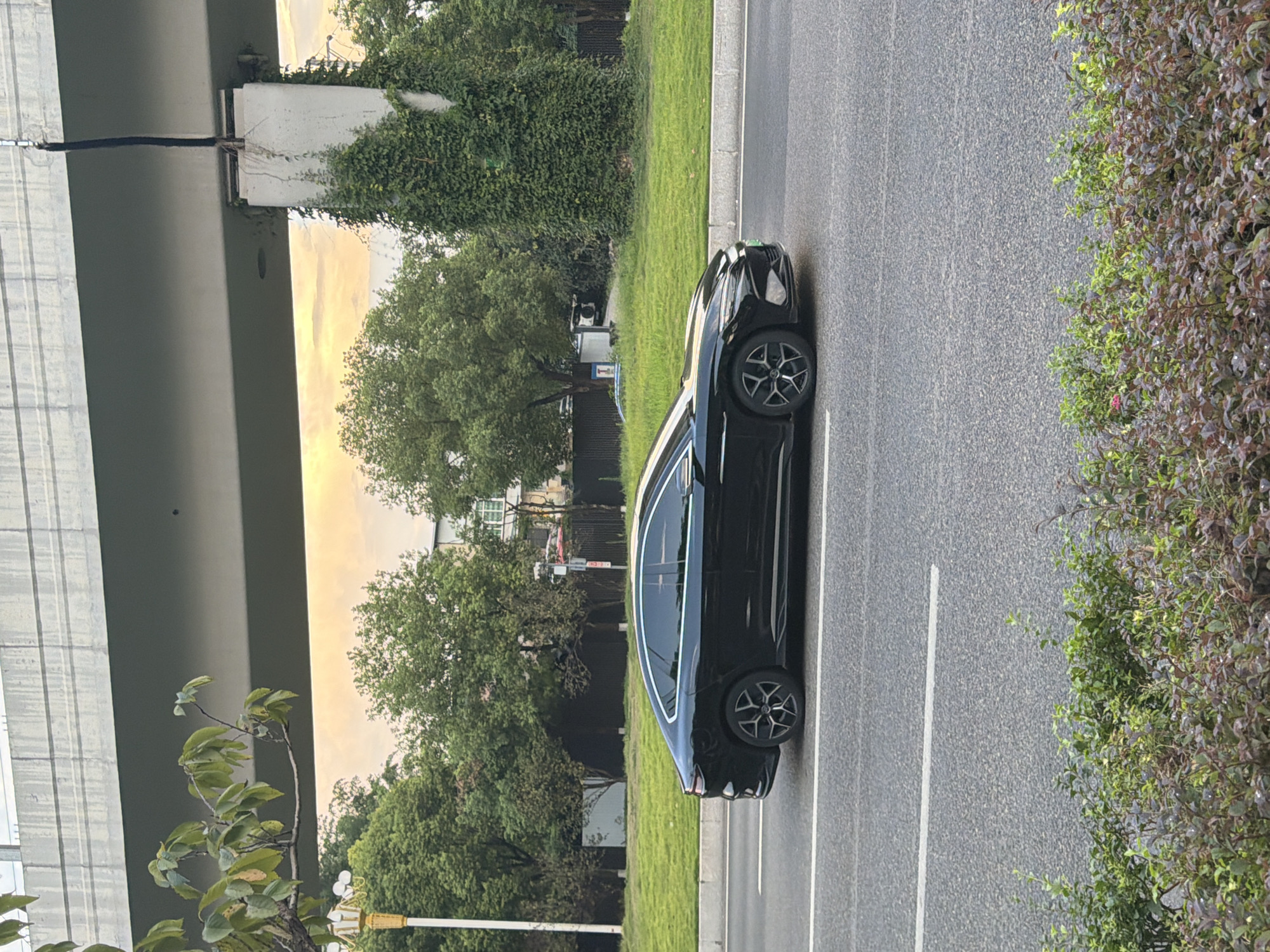 A black sedan is captured in profile, driving on a paved road in Jiangjin, China. The car is the central subject, positioned in the middle of the frame and facing to the right. Its sleek design and dark exterior are clearly visible. The road features white lane markings, suggesting it is a multi-lane street.
Behind the car, a lush green lawn stretches towards a line of mature trees. The sky above is tinged with the warm hues of sunset, indicating it is either early morning or late evening. A large, modern concrete overpass dominates the upper portion of the image, its imposing structure casting a shadow over the scene. One of the overpass's concrete support pillars is visible on the right side of the frame, partially covered in dense green ivy. To the left, a lamppost with multiple lights stands near the edge of the road. In the foreground, a dense bush with reddish-purple and green foliage adds a touch of natural detail. The overall environment suggests an urban setting with elements of both infrastructure and greenery. There are no people visible in the image.
