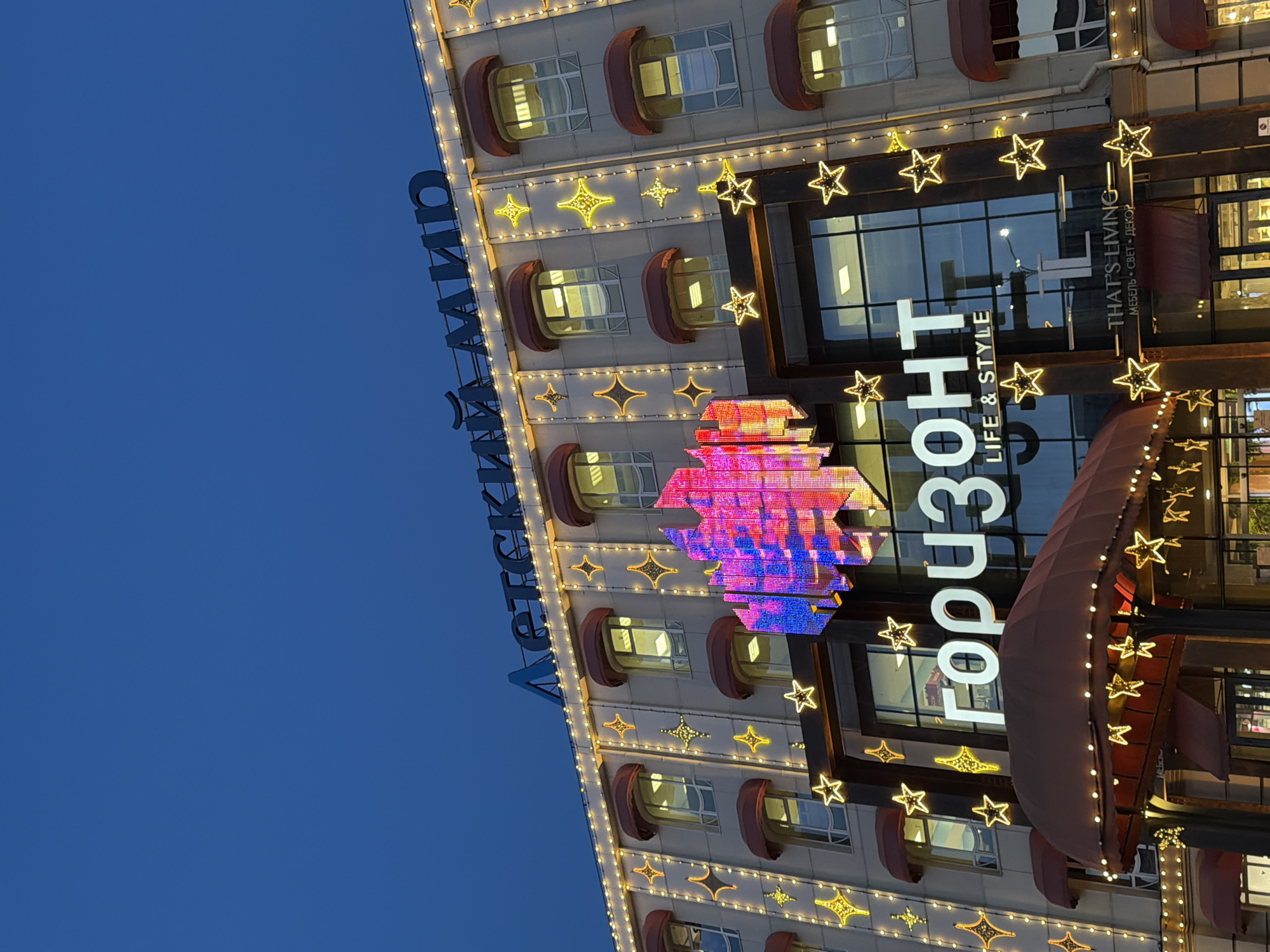 The image shows the facade of a multi-story building at twilight, likely in Rostov-on-Don, Russia. The building is decorated with illuminated stars and other decorative lighting. Two illuminated signs are visible: "Детский мир" (meaning "Children's World") vertically along the left side of the building and "ГОРИЗОНТ" (meaning "Horizon") with the tagline "LIFE & STYLE". A red and blue geometric design, possibly a logo, is above the "HORIZONT" sign. The building also features a red awning and windows with rounded, red overhangs. The sky is a deep blue.