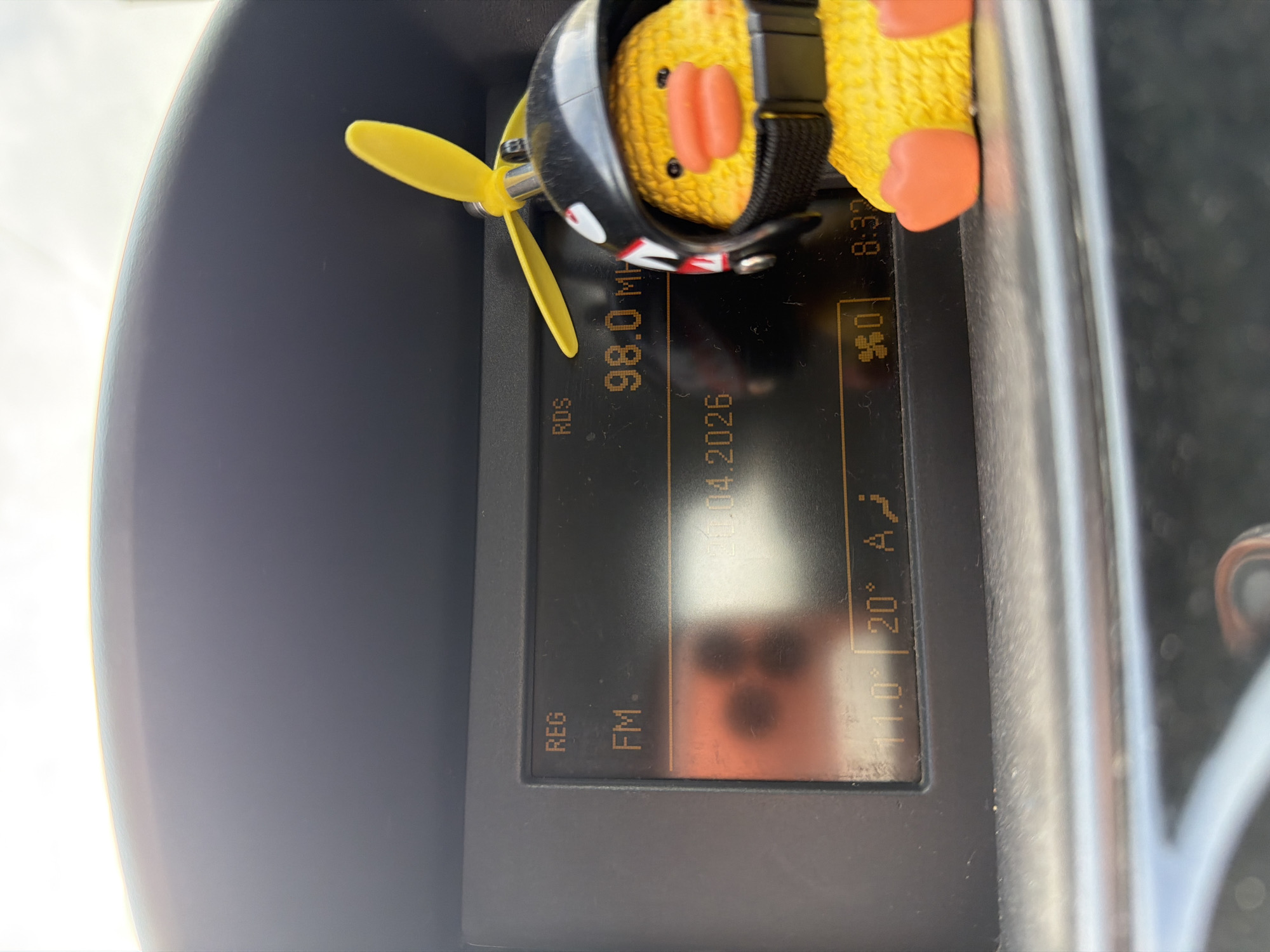 The image shows the interior of a vehicle, specifically the dashboard area, likely located in Sofia, Bulgaria (based on the Cyrillic script potentially used for the "A" in the temperature display). A yellow rubber duck wearing an aviator helmet with a propeller is resting on top of the car's infotainment screen. The infotainment screen displays radio information (FM 98.0 MHz) and indicates the date as April 4, 2026. The temperature is displayed as 11.0°C, with the air conditioning set to 20°C.