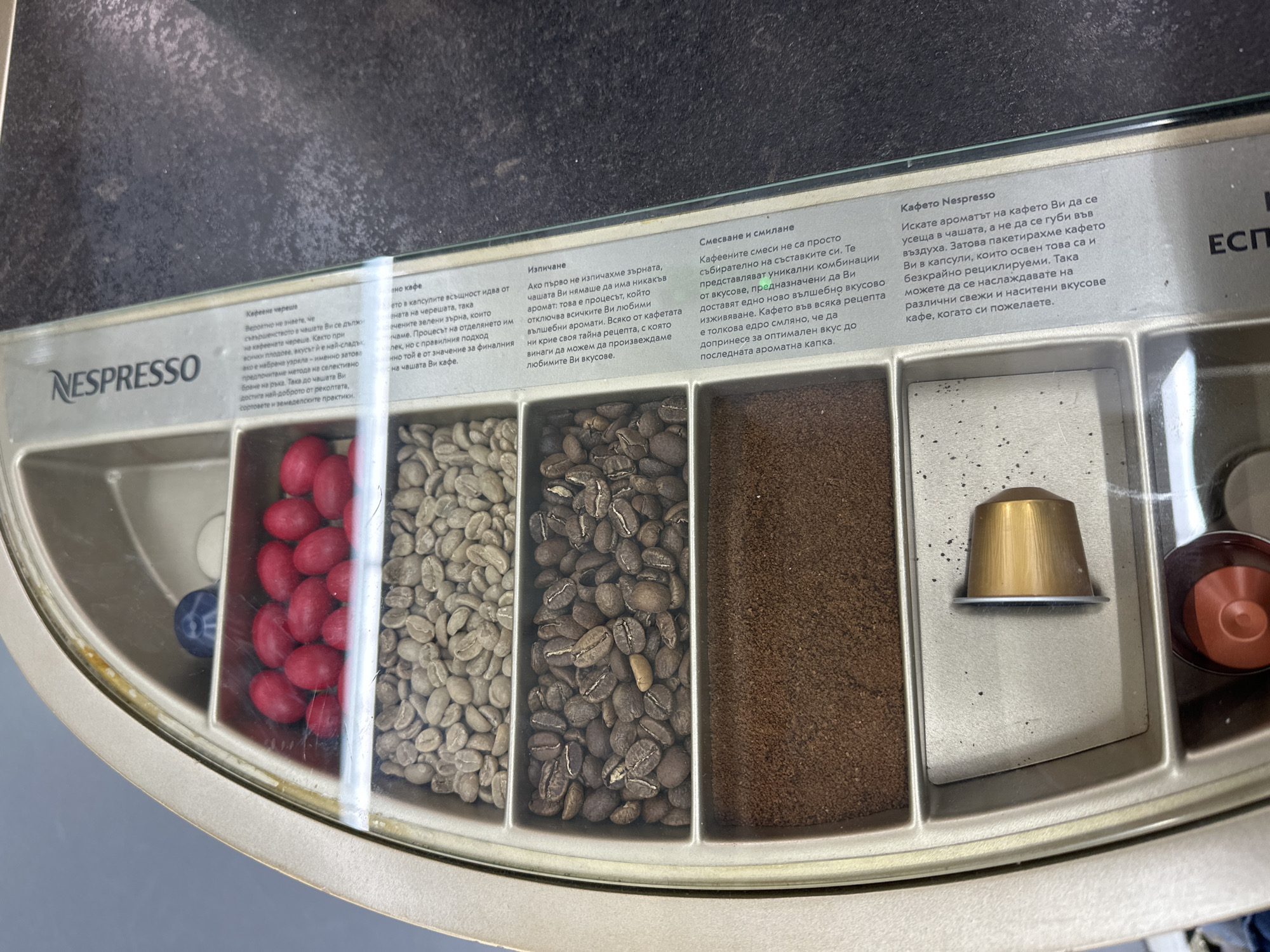A close-up, eye-level shot captures a Nespresso display case, likely found in a retail environment in Sofia, Bulgaria. The display, protected by a clear cover, illustrates the journey of coffee from bean to capsule, arranged sequentially from left to right.
On the far left, bright red coffee cherries are showcased, followed by a compartment filled with pale green, unroasted coffee beans. The next section contains dark, roasted coffee beans, and then a compartment holding finely ground coffee. The final section on the right prominently features a gold-colored Nespresso coffee capsule, with another red/brown capsule partially visible behind it.
Above each stage, informational text in Bulgarian explains the process. The first section, titled "Кафеена череша" (Coffee cherry), discusses the importance of hand-picking ripe cherries for optimal flavor. The "Изпичане" (Roasting) section details how roasting unlocks the coffee's aromatic profile. "Смесване и смилане" (Blending and grinding) explains how unique flavor combinations are achieved through precise grinding. Finally, "Кафето Nespresso" (Nespresso coffee) describes the brand's method of preserving freshness and intense flavors by packaging coffee in recyclable capsules. The Nespresso logo is clearly visible on the left, and a partially visible "ЕСП" is on the far right. The display aims to educate consumers on the quality and process behind Nespresso coffee.