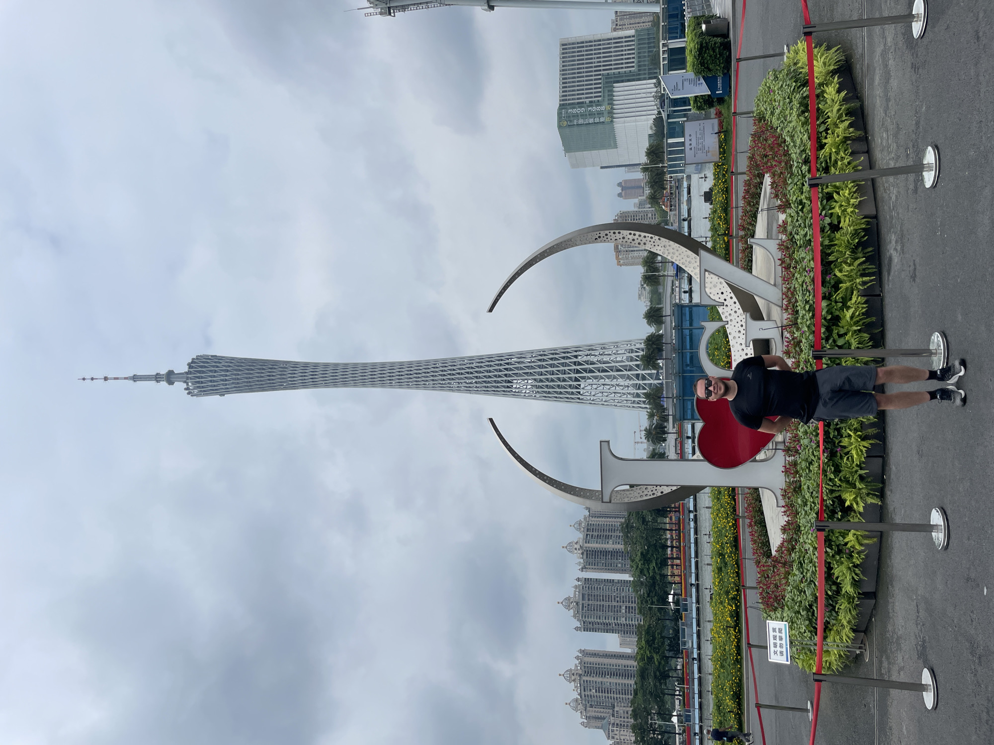 A medium-shot captures a man standing next to a large, decorative metal structure featuring a heart, with the Canton Tower prominently visible in the background, under a cloudy sky in Tianhe District, China. The man, wearing a black t-shirt, dark shorts, sunglasses, and athletic shoes, stands to the right of the metal sculpture. A line of potted plants with green and red foliage separates the structure from the pavement. Red rope and metal stanchions guide visitors around the area. Behind the sculpture and the man rises the Canton Tower, a towering lattice structure reaching into the sky. Numerous buildings of varying heights populate the distant skyline behind the tower. The sky is overcast with a uniform gray color.