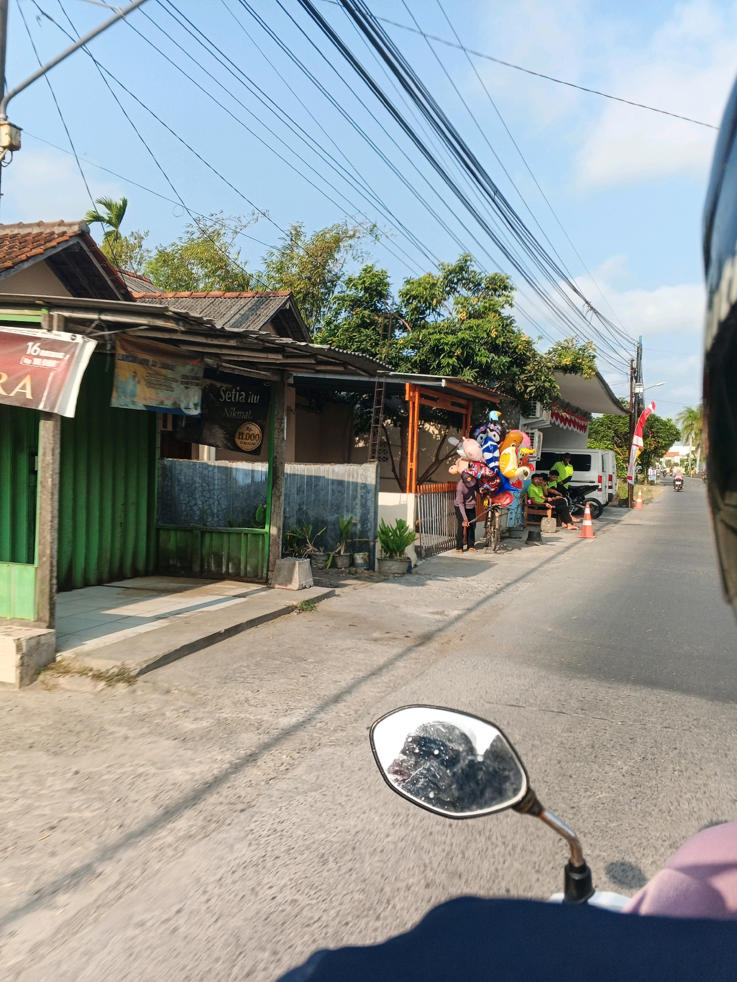 This is a medium-distance, eye-level, exterior shot taken from a moving motorcycle, looking forward and slightly to the left. The scene depicts a street in a residential or commercial area with a clear blue sky above, crisscrossed by numerous overhead utility wires. On the left side of the street are several buildings with tiled roofs and corrugated metal or green siding. One building has a sign that says "Setia itu Nikmat" with a price of Rp. 11.000. In front of this building, a person is standing next to a bicycle laden with colorful balloons, including some shaped like animals. Further down the street on the right, there are several orange traffic cones, a white van, and more buildings. Three young men are visible sitting on a bench near the van. Other pedestrians and a motorcycle are seen in the distance on the right side of the road. The street itself is paved with asphalt. The foreground includes the motorcycle's side mirror, which is reflecting the rider's helmet and the sky, and a portion of the motorcycle's seat. The overall impression is a snapshot of everyday life on a street in what appears to be Southeast Asia.