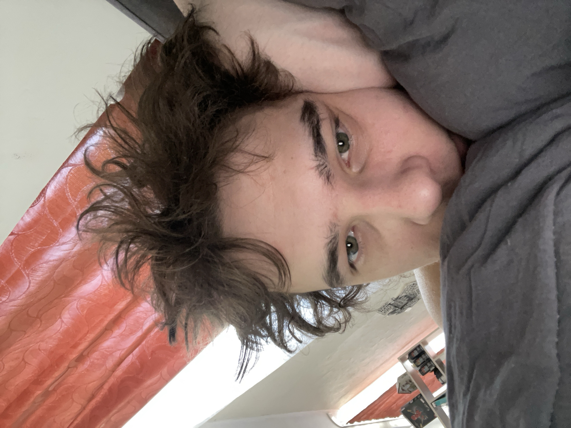 The media file is an image, rotated 90 degrees clockwise, depicting a close-up portrait of a person. The subject, likely a young adult, is shown from an overhead perspective, looking directly into the camera while seemingly lying down. They have messy, dark brown hair and light skin. Their left eye, featuring a greenish-hazel iris, is clearly visible, along with part of their nose and forehead. A dark grey blanket or duvet covers a significant portion of their body and the right side of the frame (when upright). To the left, an orange or reddish curtain with a subtle patterned texture is visible. Above the curtain and extending across the upper part of the frame is a plain, light-colored wall or ceiling. In the blurred background, on the lower right (when upright), some indistinct furniture or shelving with objects can be seen. The person's expression is neutral, suggesting a relaxed or possibly just-woken state, emphasized by the disheveled hair. The overall atmosphere suggests an indoor, relaxed setting, possibly a bedroom. No discernible text is visible in the image. This scene is located in Krasnoyarsk, Russia.