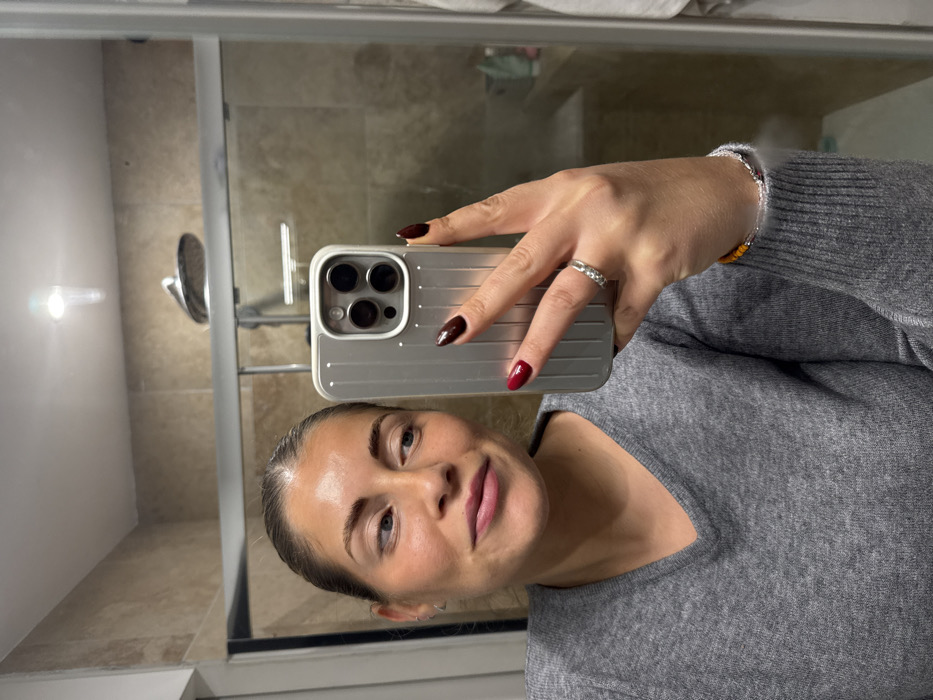 A woman takes a selfie in a bathroom mirror. She wears a gray sweater and a ring on her left ring finger. Her nails are painted dark red and maroon. The phone in her hand has a silver, ridged case. A showerhead is visible in the mirror behind her. The bathroom has tiled walls and a light fixture.