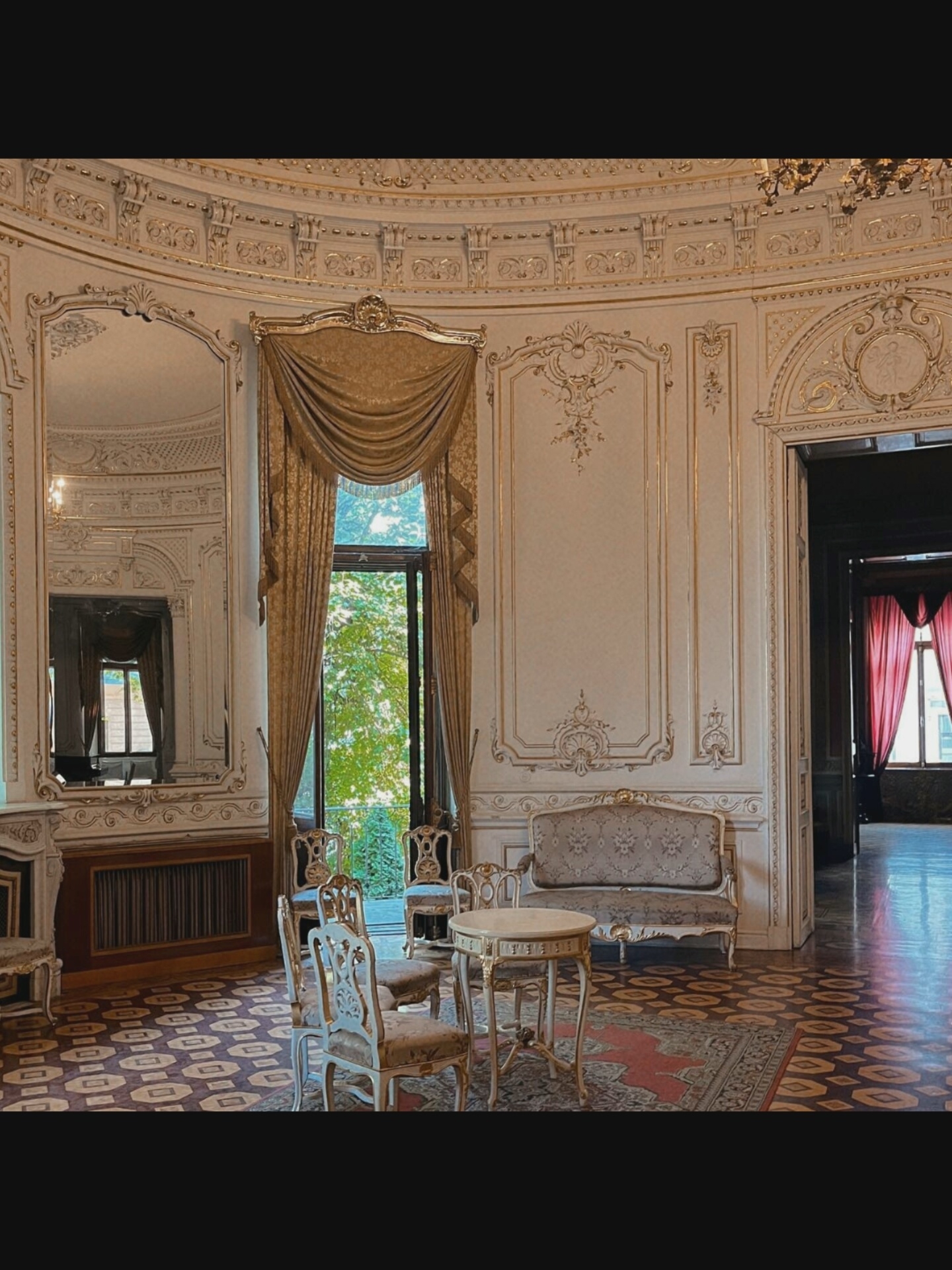 The image depicts an opulent, ornate interior room, likely a reception or drawing-room, characterized by lavish gold detailing on white walls and ceilings, intricate moldings, and a large, decorative mirror on the left. The room features parquet flooring with a geometric pattern, a floral-patterned rug, and a collection of antique furniture.
In the center of the room, a small, round table with gold accents sits surrounded by four ornate chairs, two of which are visible in the foreground. To the right of the table is a small, two-seater sofa with similar decorative upholstery.
A large, arched window or doorway, draped with heavy golden fabric, is visible in the background, offering a view of lush green foliage and a balcony railing. To the far right, a dark doorway reveals another room with deep red curtains and a polished floor, suggesting a connected space.
The overall impression is one of grandeur and historical significance, possibly a salon in a historic building or a museum. The specific mention of Saint-Août, France, suggests this room is located within that geographical area. There are no people visible in the scene, and no actions are being performed. The scene is static and focused on the architectural and decorative elements of the room.