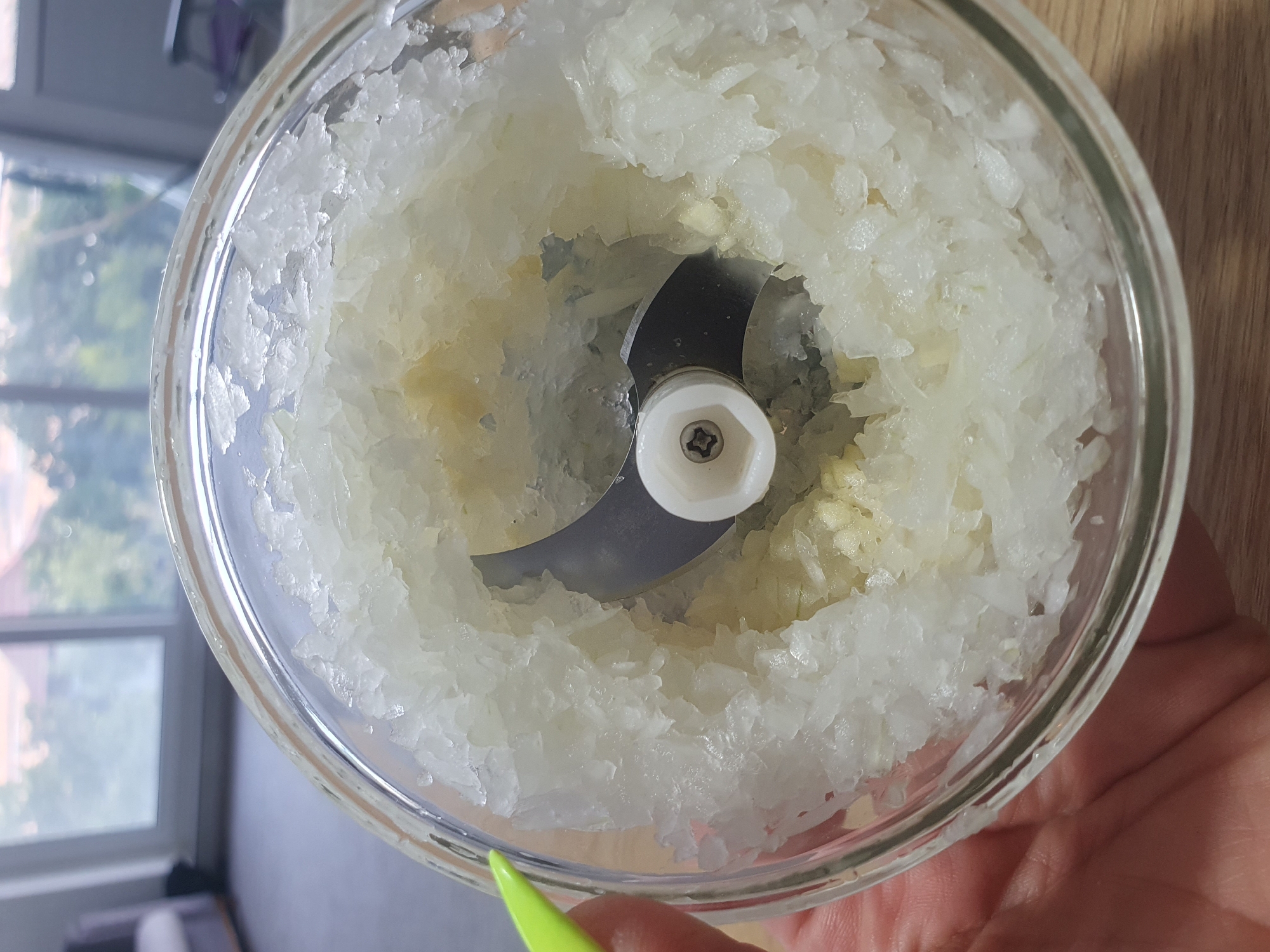 A close-up shot shows the interior of a glass food chopper filled with finely minced white onion. A single silver blade is visible at the center, attached to a white plastic mechanism. The minced onion surrounds the blade and extends to the edges of the glass container. A person's hand, with a neon green fingernail, is visible on the left side of the frame, holding the chopper. The background is out of focus and appears to be an indoor setting with a window showing greenery and a glimpse of distant buildings, possibly indicating a residential area. There is no information in the image to suggest the location is Kheyrabad, Iran.