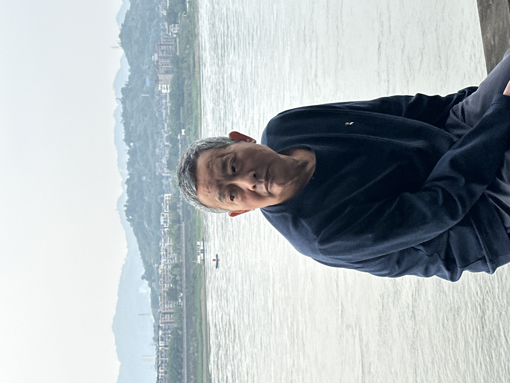 A man is sitting in front of the Yangtze River in the city of Jiangjin, China. The man is gray-haired and wearing a dark sweater. The river is wide and gray-green. Buildings and mountains are visible on the far bank of the river. A small boat is on the river.