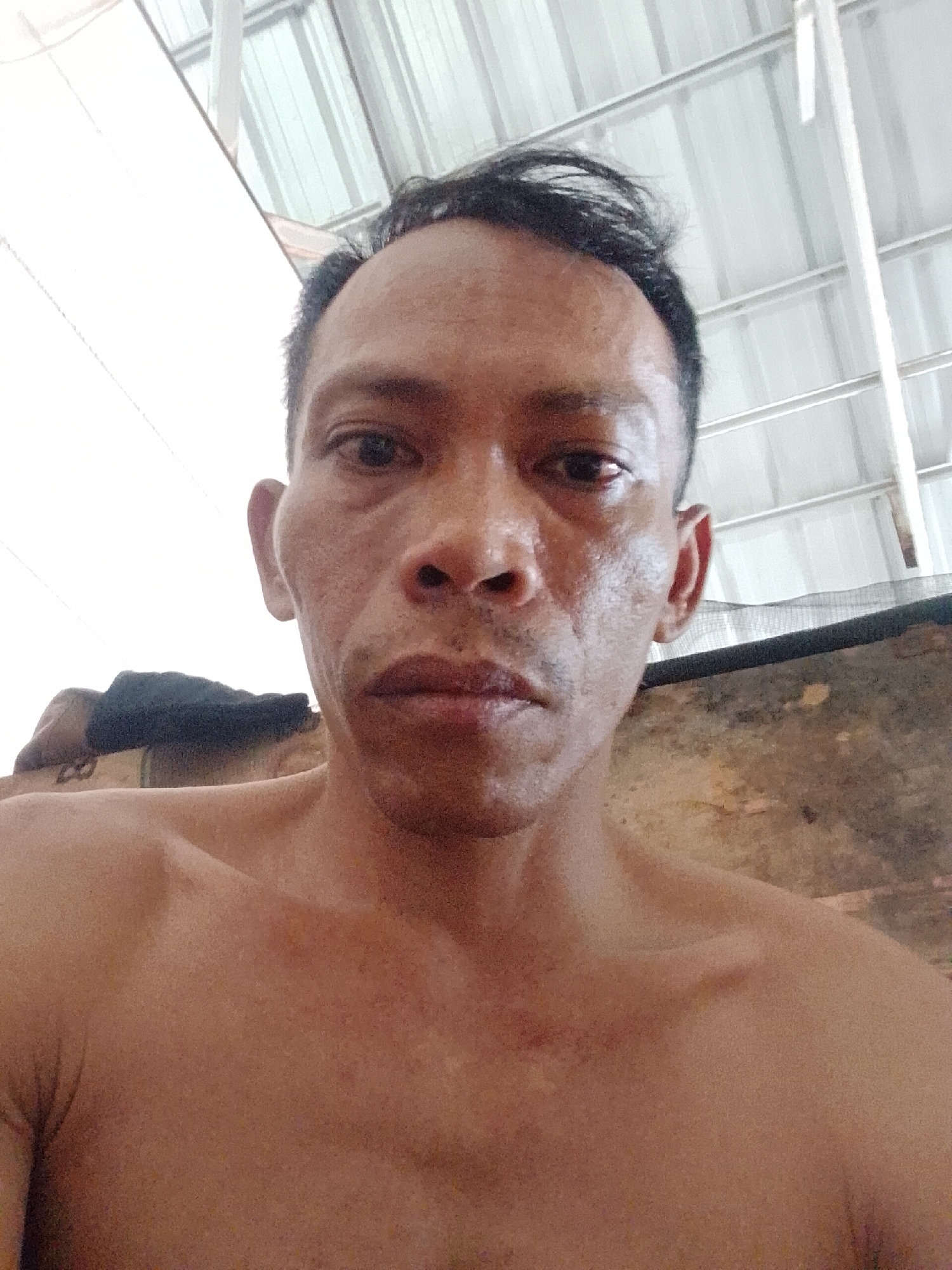 A close-up, underexposed photograph shows a bare-chested, middle-aged man with dark hair and a receding hairline. His skin shows signs of sun damage. He is looking directly at the camera with a neutral expression. Behind him is a corrugated metal roof. To the left of the man is an indistinct object covered in black material. The image was taken in Bukit Ketok, Indonesia.