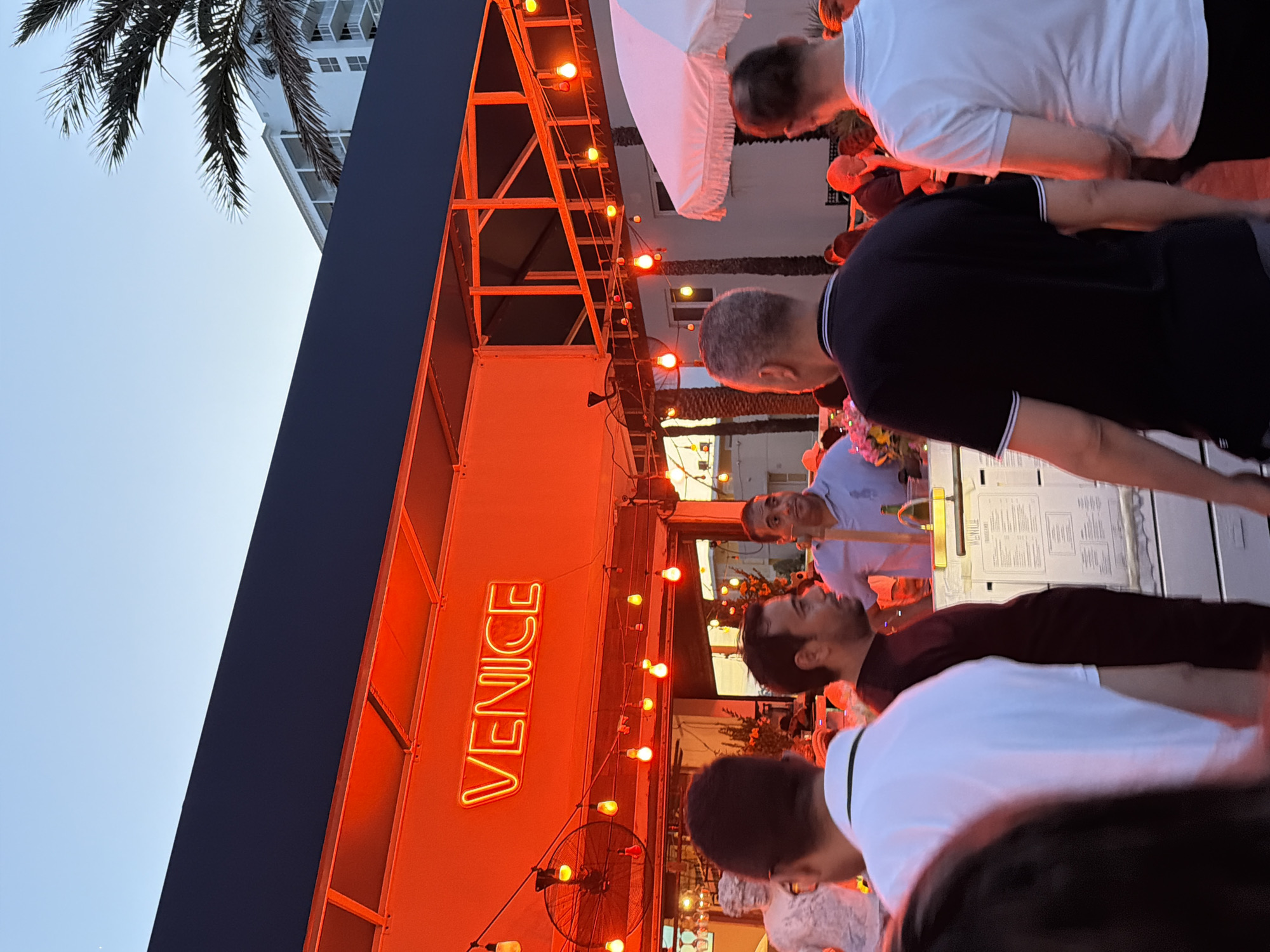 The image depicts an outdoor scene at dusk, likely in the city of Hollywood, United States of America, with a prominent neon sign reading "VENICE" glowing orange. The setting appears to be a restaurant or bar with an open-air setup, indicated by the strung lights and the awning. Several people are visible, engaged in conversations and milling about. In the foreground, several individuals are seen from behind, some wearing casual attire like t-shirts. A white statue, possibly a bust, is partially visible on the left. Behind the counter, a man in a light blue shirt is interacting with someone. The overall atmosphere suggests a lively evening gathering.