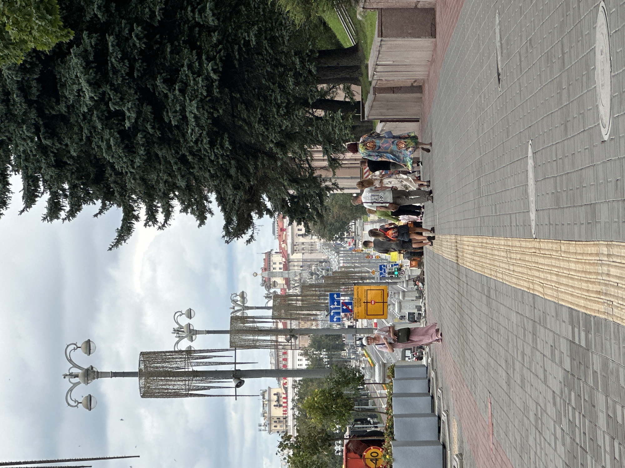A medium shot captures a street scene in Minsk, Belarus, on a cloudy day. The foreground is dominated by a paved walkway made of gray cobblestones, with a tactile paving strip of yellow concrete tiles running down the center. The walkway is lined with planters filled with greenery.
In the middle ground, a group of people are walking away from the camera. A woman in a long pink dress is walking on the left side of the walkway. To her right, several other individuals are walking in the same direction, including a man in a white t-shirt, a woman in a floral dress, and others.
Further down the street, there are decorative streetlights with ornate metalwork and globes. A large evergreen tree stands on the right side of the frame, its dark green foliage providing a strong contrast to the lighter sky. In the background, multi-story buildings with classical architecture line the street. Traffic and street signs are visible, including a yellow sign with Cyrillic text and arrows, and a speed limit sign displaying "40". The overall impression is of a well-maintained urban environment.