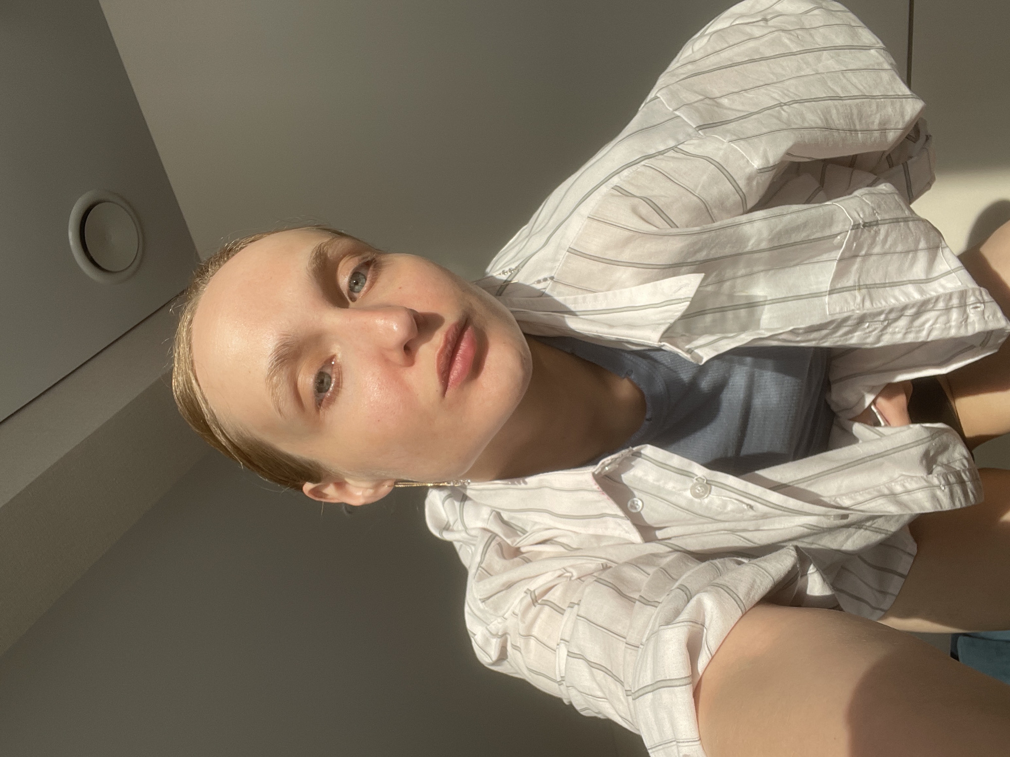 A selfie image features a light-skinned person wearing a gray-blue top and a white shirt with light gray vertical stripes. The subject is positioned at an angle, looking upwards towards the camera. The background consists of a plain wall, with a circular air vent visible in the upper left corner. The image is well-lit, with natural sunlight casting shadows on the wall and the subject's face and clothing. Based on the person's appearance and the style of the image, it is likely a recent photograph taken in an indoor location. It is impossible to determine if the image was taken in Sepych, Russia, or any other specific location based on the visual information alone.