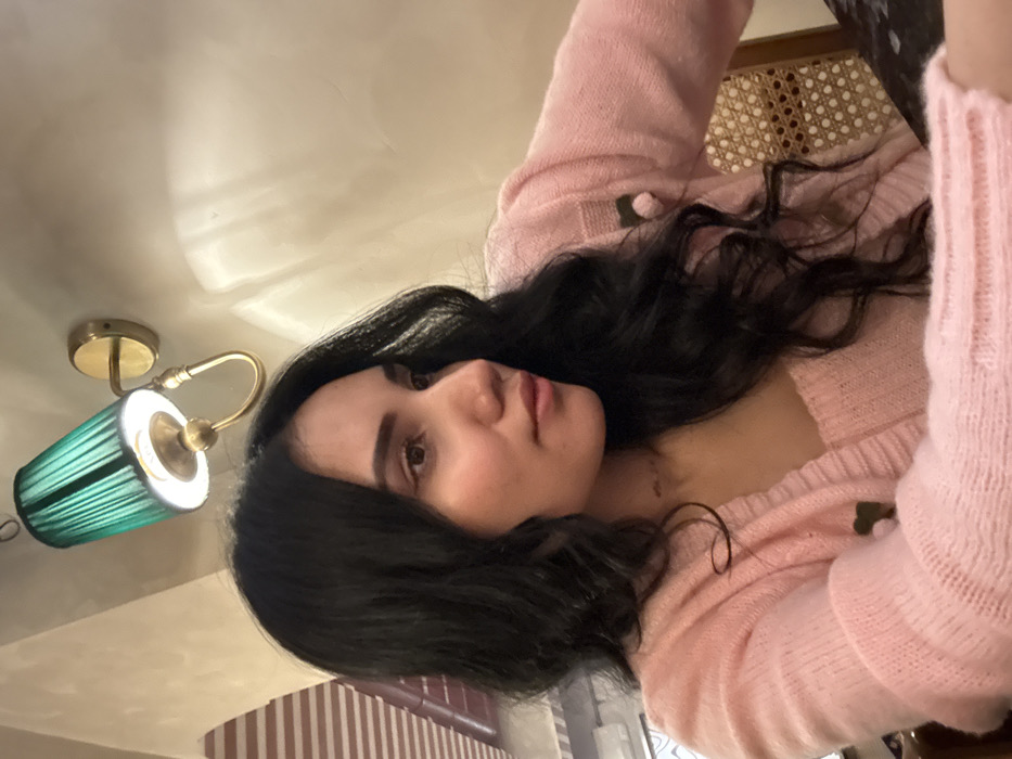 An indoor scene features a female individual, observed from the chest up, positioned slightly to the left of the frame's center. She possesses long, dark, wavy hair and an olive skin tone, with her gaze directed towards the right. She is attired in a light pink knitted cardigan worn over a matching light pink knitted top. A thin necklace is visible around her neck.

In the upper left background, a wall-mounted sconce with a brass-toned fixture and a pleated dark green fabric shade is illuminated, casting light on the surrounding light-colored wall. To the subject's right, the woven cane backrest of a chair is partially visible. The background wall exhibits subtle light and shadow variations. A faint, striped pattern is discernible in the far upper left background. The specific location within the city of Howrah, India, cannot be ascertained from the visual evidence presented in the image. No other individuals or direct interactions are depicted.