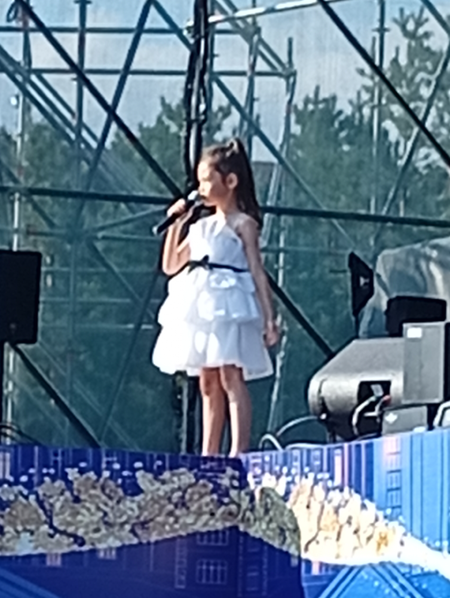 A young girl with dark hair in a ponytail, wearing a white tiered dress and holding a microphone, stands on a stage. She is positioned towards the right side of the frame, looking to her left. In the background, a metal scaffolding structure is visible against a bright sky, with trees behind it. To the left of the girl, a black speaker is mounted on a pole. To her right, a silver piece of equipment with black accents is situated on the stage. The stage itself is decorated with a blue, patterned backdrop that appears to depict buildings and textured elements. The image quality is somewhat blurry. The location is identified as Kostanay, Kazakhstan.