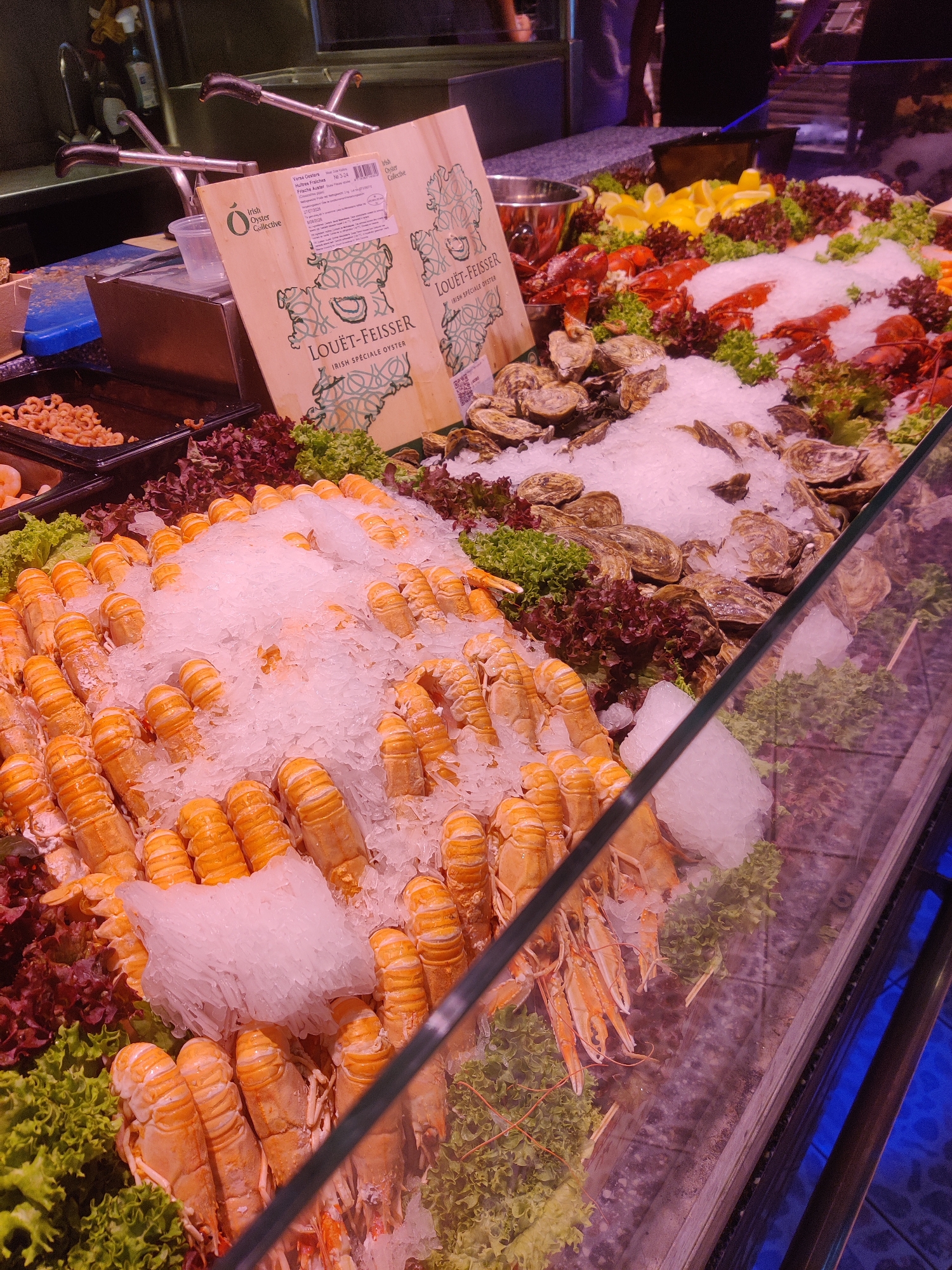 The image depicts a seafood display at a market, likely in The Hague, Netherlands. The primary focus is on a generous arrangement of seafood, primarily langoustines and lobsters, served on crushed ice. The langoustines are positioned in the foreground, meticulously laid out on a bed of ice, with their bright orange bodies and segmented tails clearly visible. Behind them, a collection of lobsters, also on ice, are arranged with their red shells contrasting against the white ice.
Scattered amongst the seafood are clusters of oysters, their rough shells nestled in the ice. Lemons, sliced into wedges, and various types of lettuce, including red leaf and frilly green varieties, are interspersed throughout the display, adding color and freshness.
Two wooden signs with intricate green swirling patterns, possibly representing maps or decorative elements, are propped up behind the seafood. Text on these signs, partially obscured, reads "LOUËT-FEISSER" and "IRISH SPÉCIALE OYSTER." One sign also has a white label with smaller text, indicating it is from the "Irish Oyster Collective."
In the background, metal taps and plumbing fixtures are visible, suggesting a preparation or washing area. A dark bin containing small, pinkish seafood, possibly shrimp or prawns, is also present. The overall environment appears to be a well-lit indoor market stall, with a glass counter separating the viewer from the display. The lighting casts a slight pinkish hue over the scene. No individuals are clearly visible interacting with the display, though the presence of market infrastructure implies a commercial setting.