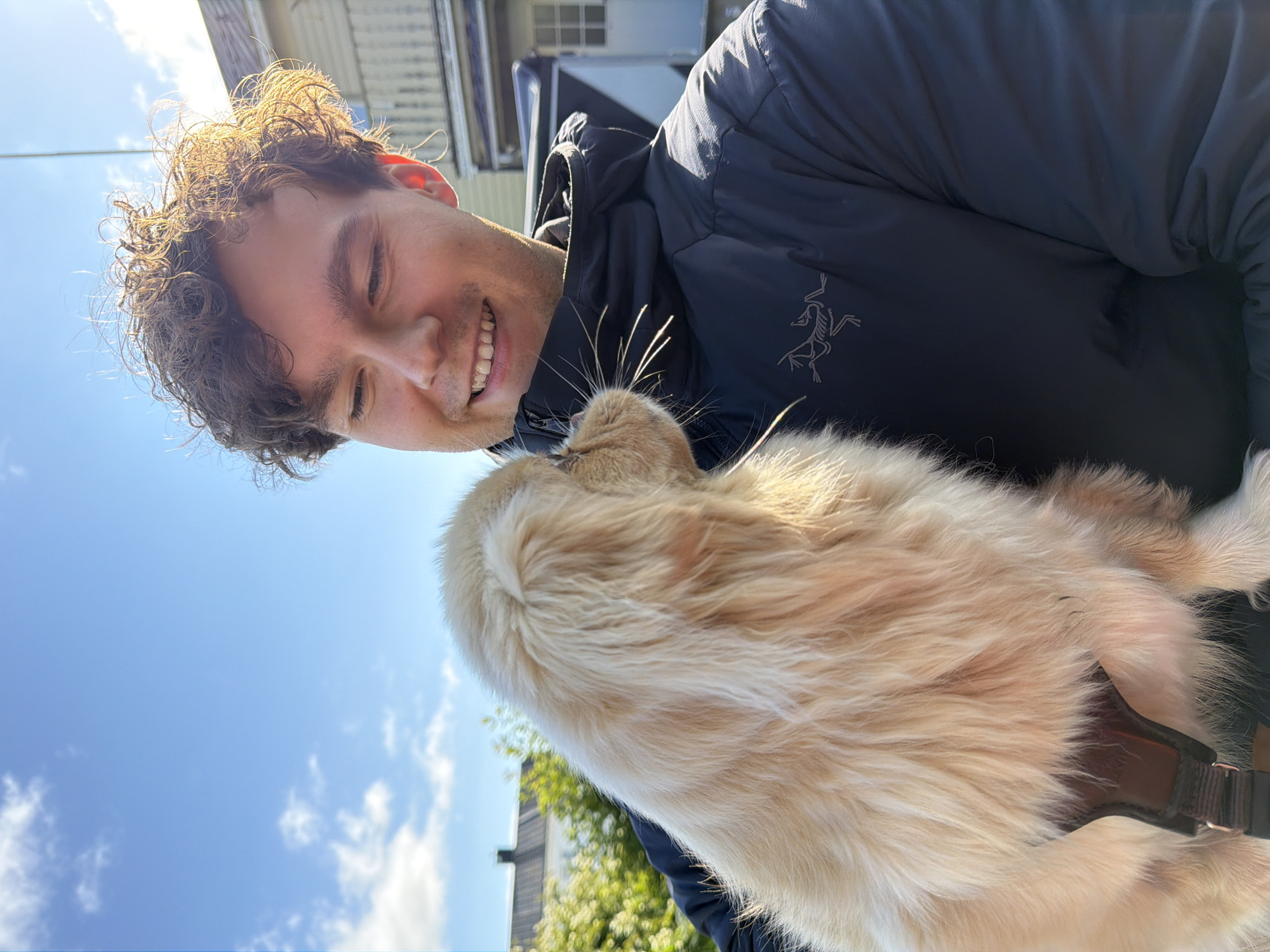 A young man with curly brown hair, wearing a dark blue puffer jacket with an Arc'teryx logo on the left chest, is holding a golden-colored puppy. The man is smiling and looking down at the puppy. The puppy is facing away from the camera and towards the man. The background shows a blue sky with some clouds, and buildings that appear to be residential houses. The image was taken outdoors, likely on a sunny day. The scene appears to be in a residential area.