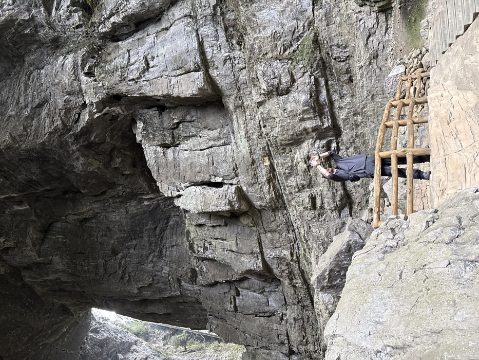 The image displays a male individual standing within the Three Natural Bridges (Tiankeng Three Bridges) karst geological formations in Wulong District, Chongqing, China. The individual, dressed in a dark grey short-sleeved shirt, dark trousers, and dark shoes, stands on a light-colored paved pathway secured by a light brown wooden railing. He is holding a white smartphone or small camera with both hands, aiming it upwards, seemingly photographing the towering grey rock walls. A large natural archway is visible in the upper left of the frame, revealing a brighter, vegetated exterior beyond. The surrounding environment consists of immense, stratified grey rock formations, characteristic of a deep natural gorge or cave system.