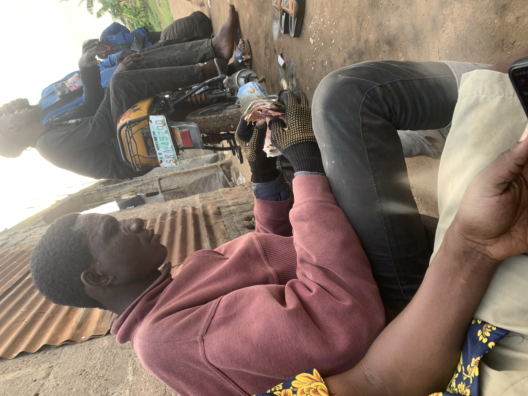 A man is sitting on the ground in a dusty outdoor area, wearing a maroon sweatshirt and gray pants. He has on black gloves with yellow dots and is holding a wad of Nigerian Naira banknotes. Behind him, a yellow motorcycle is parked, with two other men sitting on it. The man on the motorcycle in the foreground wears a black long-sleeved shirt and dark pants. The man behind him wears a blue long-sleeved shirt. Another man wearing a blue jacket is standing in the background. The scene appears to be in Shagamu, Nigeria, with a corrugated metal wall to the left and vegetation visible in the distance. The ground is dirt and dusty. The overall impression is a casual, possibly commercial, interaction.