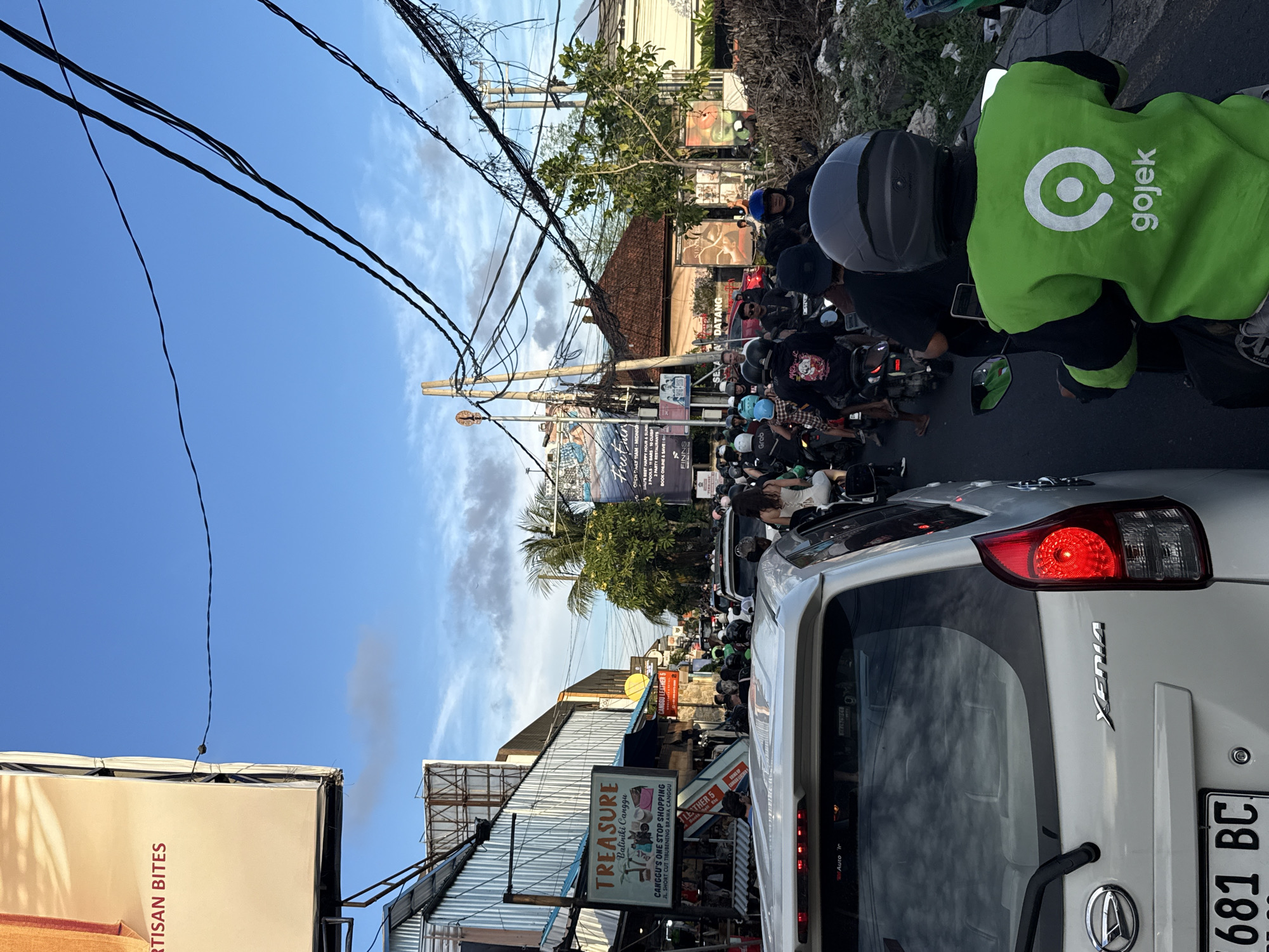 The image shows a congested street scene in North Kuta, Indonesia. Traffic is heavy, with a mix of motorcycles and cars. A Gojek driver in a bright green jacket is visible on a motorcycle. Several buildings and businesses line the street, including "TREASURE Baliniki Canggu" and a sign for "FINNS". Overhead are a tangle of power lines. The sky is blue with scattered clouds. The rear of a white car, labeled "Xenia" and with license plate "681 BC", is prominently featured in the foreground.