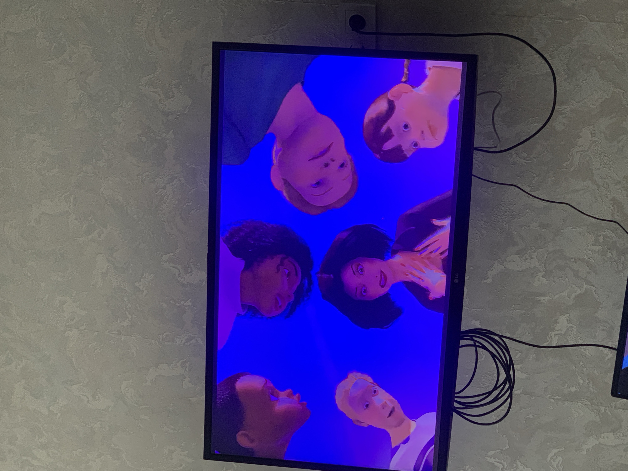 The image shows a television screen displaying animated characters. Six characters are visible, arranged in a circular pattern and appearing to look down or up at each other. The characters have stylized features, with large eyes and simplified facial structures. They are rendered in a bright, almost neon color palette, with a dominant blue background. Some characters have fair skin and light hair, while others have darker skin tones and dark hair. Their clothing is also simplified, with basic colors and designs.
The television is a modern flat-screen monitor, positioned on a wall. The wall itself has a textured, light gray finish, consistent with interior walls. Cables are visible behind the television, coiled on the wall. One cable is black and appears to be an HDMI or power cord. Another small black object is mounted on the wall above and to the left of the television, possibly a light switch or an electrical outlet. The lighting in the room appears to be artificial, as indicated by the consistent illumination of the screen and the surrounding wall.
The scene depicted on the television screen has an abstract or surreal quality due to the bright colors and the arrangement of the characters. There is no clear indication of time of day, weather, or a specific real-world location within the image itself, other than the television display. The expressions on the characters' faces are neutral or slightly curious. There is no visible text in the image. The overall impression is of a paused scene from a cartoon or animated show.
