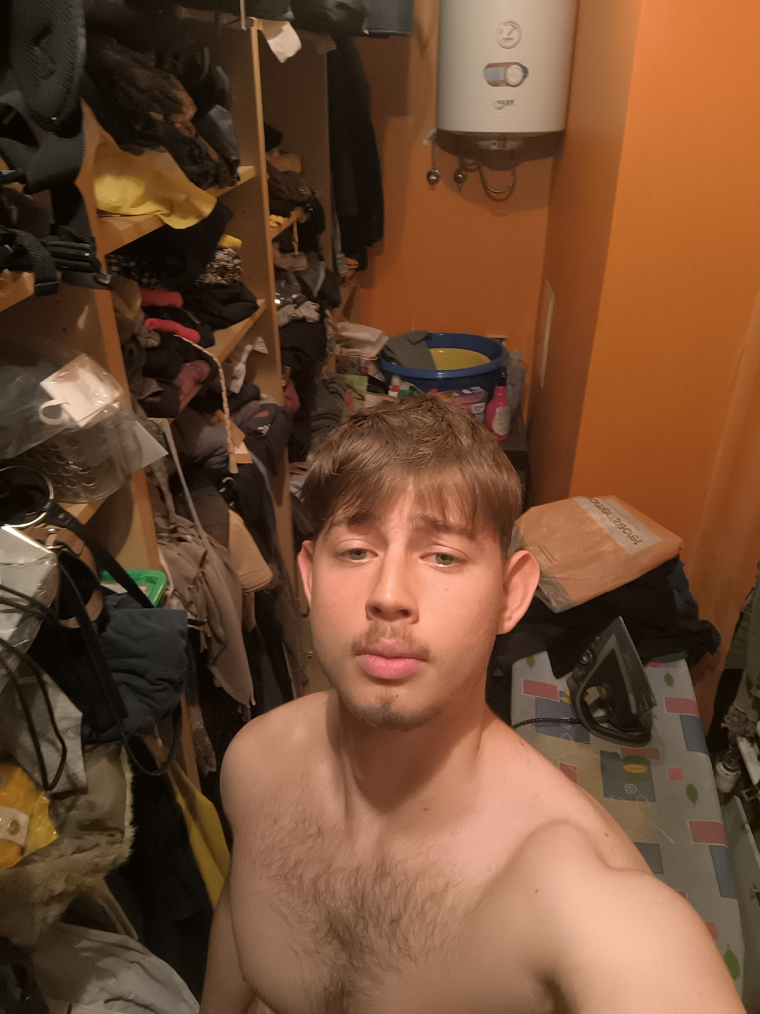 A young man with light brown hair, a mustache, and a goatee takes a shirtless selfie in a cluttered indoor room. His expression is neutral as he looks directly into the camera. The room is characterized by bright orange walls and artificial lighting, suggesting evening or night.
To the left, a tall, narrow wooden shelving unit is overflowing with clothes, bags, and various household items. In the upper right corner, a white electric water heater or boiler, featuring a "Z" logo and control dials, is mounted on the orange wall. Below the water heater, a blue bucket with a yellow insert contains some unspecified items. On the right side of the frame, a dark iron rests on an ironing board with a patterned cover in grey, blue, and pink. Behind the iron, a brown package or box is visible, stacked on top of clothes, displaying reversed text that appears to read "emomew.eL".
The overall scene conveys a domestic setting, possibly a utility room, storage area, or a small room within an apartment in Sofia, Bulgaria, given the provided context. The space is visibly unorganized, with many personal belongings crammed together.