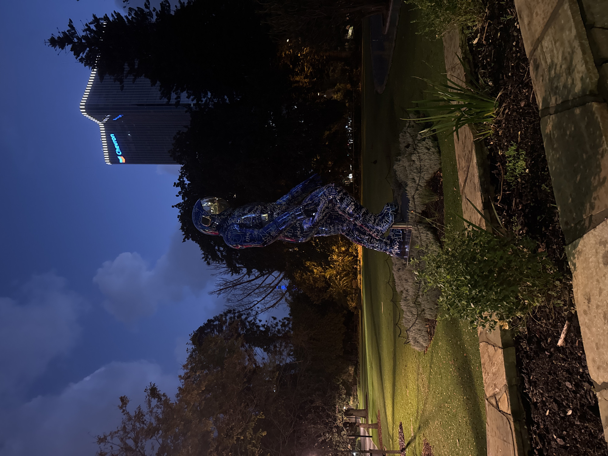 The image shows a nighttime view in Perth, Australia, featuring a distinctive sculpture of a person wearing a helmet, lying prone. The sculpture is dark, with blue and white illuminated elements, potentially made of a metal frame covered with patterned material.
The Chevron building is visible in the background, its illuminated signage indicating its identity. Trees frame the composition, and a small area of grass and a raised garden bed are visible near the sculpture. A paved walkway and part of a bench can be seen in the lower portion of the frame. The sky is a deep blue, with clouds visible. The lighting suggests it's evening. The scene appears to be taken within a park or garden setting close to Perth's Central Business District.