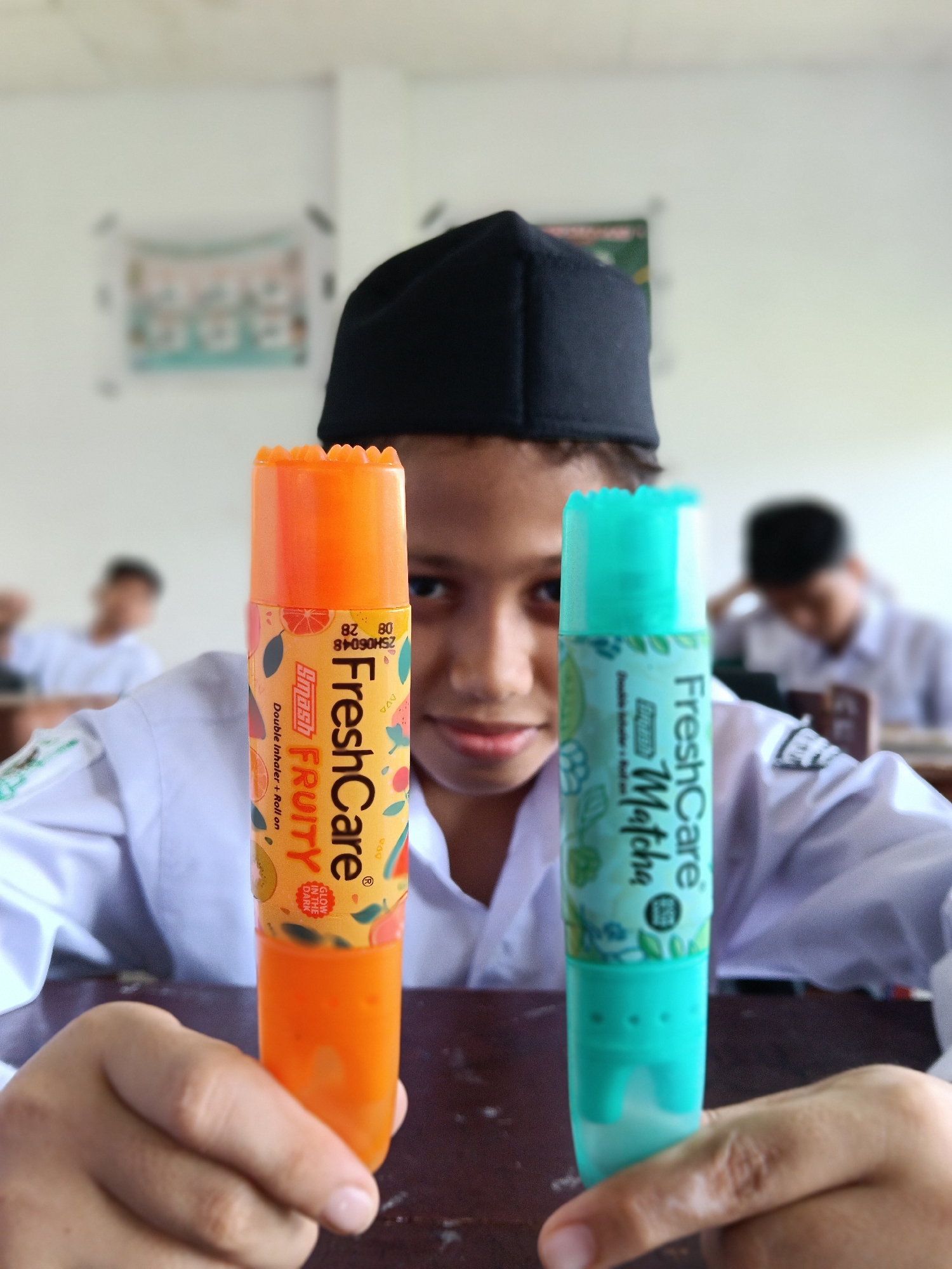 In a classroom setting, an individual, presumably a student, is showcasing two FreshCare aromatherapy inhalers. The one in the left hand is an orange 'Fruity' scent, while the one in the right hand is a light blue 'Matcha' scent. The subject, wearing a white shirt and a black peci hat, is sitting at a desk. Other students can be seen blurred in the background. The image was taken in the city of Lhokseumawe, Indonesia.