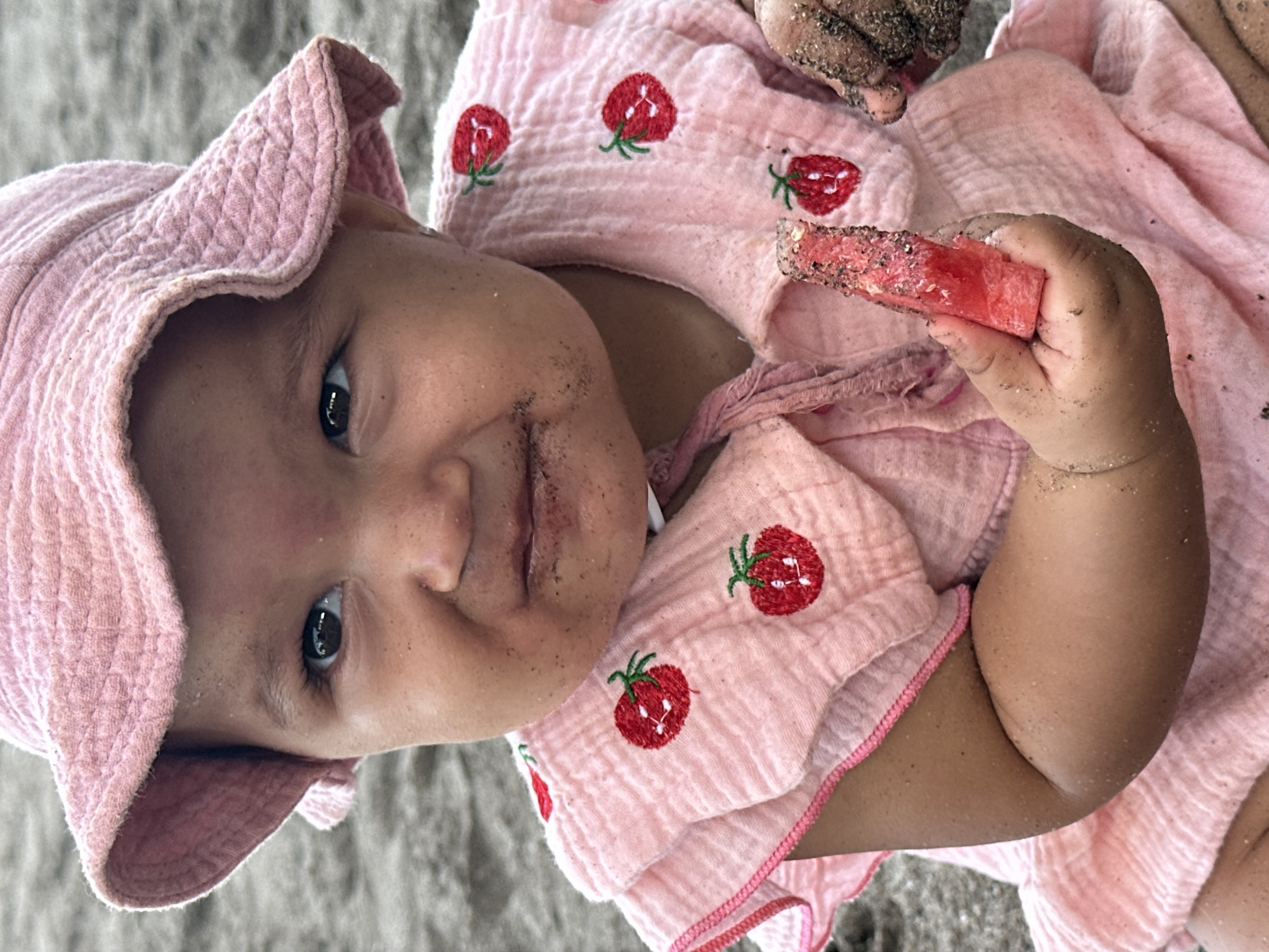 A young child wearing a pink sun hat and a pink outfit adorned with strawberry embroidery is holding a piece of watermelon. The child's face and hands are lightly covered in sand. The child is on a sandy surface which may be the beach in North Kuta, Indonesia.