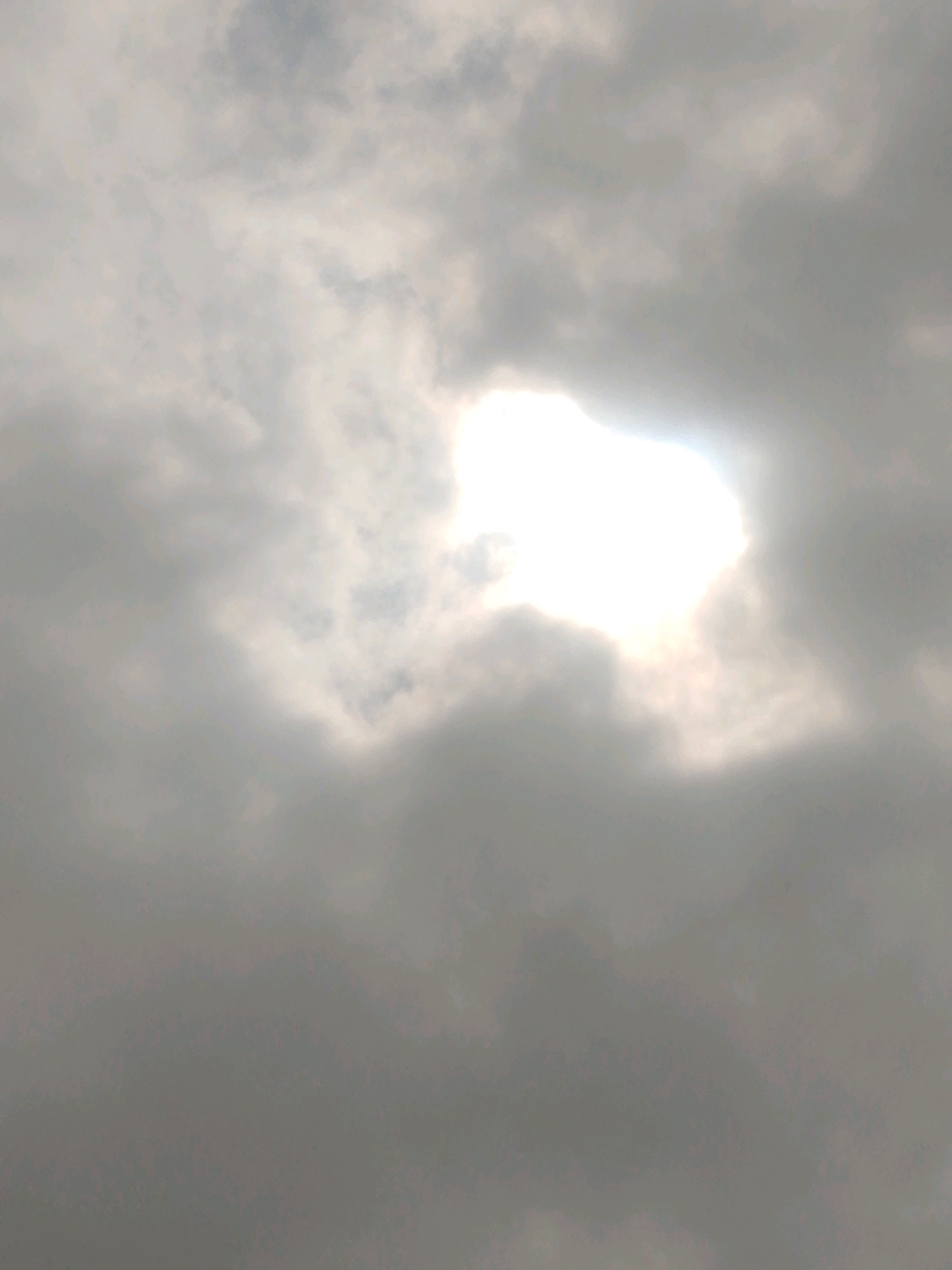 The image shows a sky with clouds. The sun is visible through a break in the clouds, creating a bright, radiant area. The clouds are light gray and white, with darker gray areas in some places. There are no visible buildings, people, or other objects in the image, suggesting it is a photograph of the sky. The location in Masaka, Nigeria, is not discernible from the image itself.